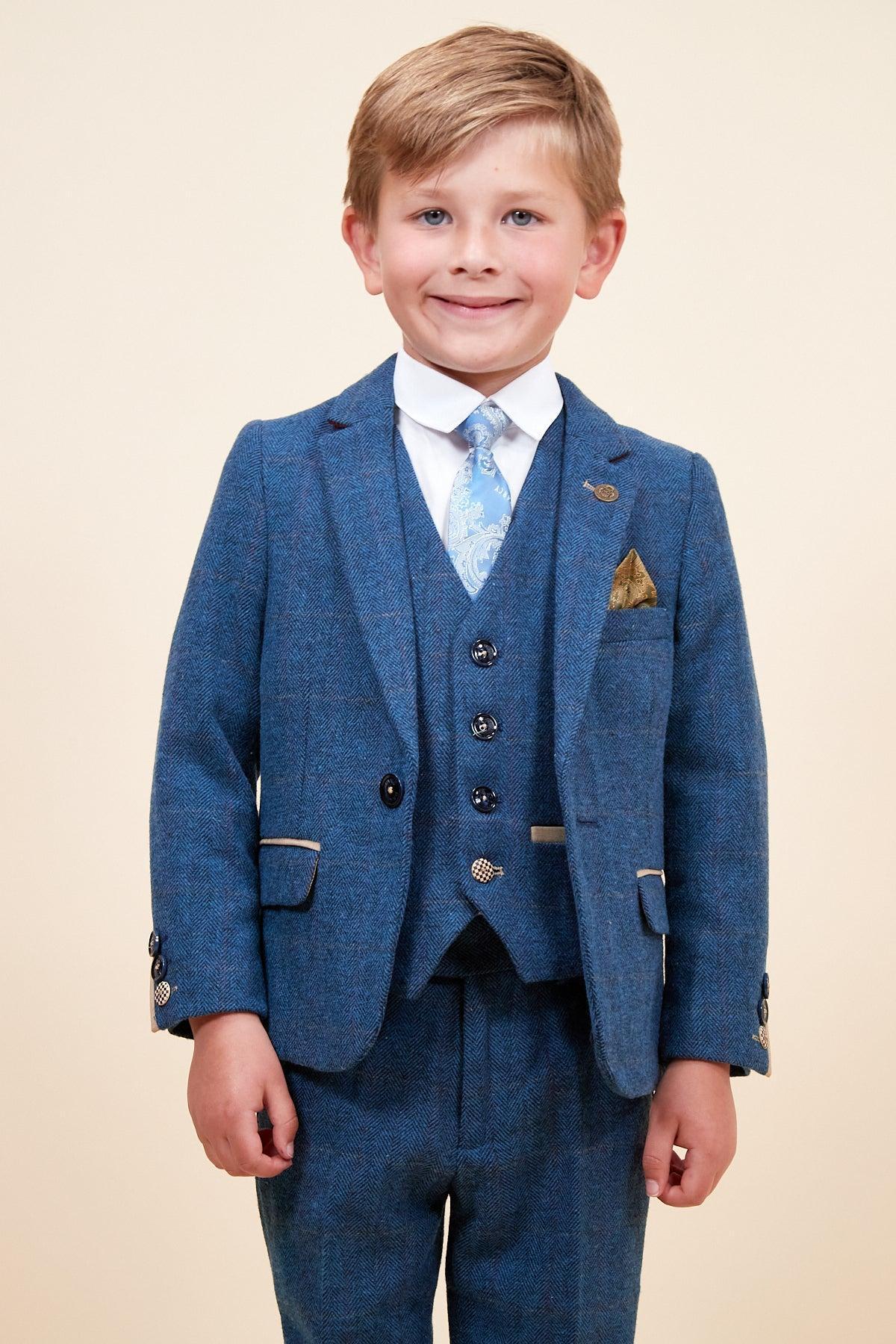 A blonde boy smiles in the marcdarcy DION - Childrens Blue Tweed Check Three Piece Suit, featuring a matching vest, white shirt, patterned tie, and gold pocket square. He stands against a plain beige background.