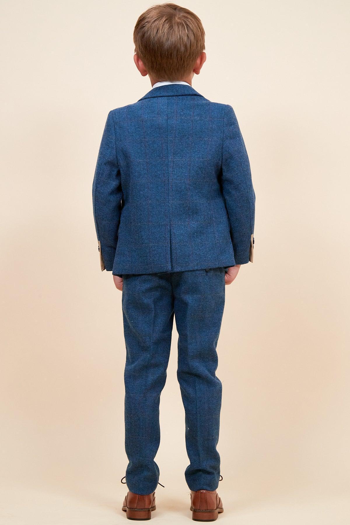 A young boy faces away from the camera wearing the marcdarcy DION Childrens Blue Tweed Check Three Piece Suit with brown shoes, set against a plain beige background.
