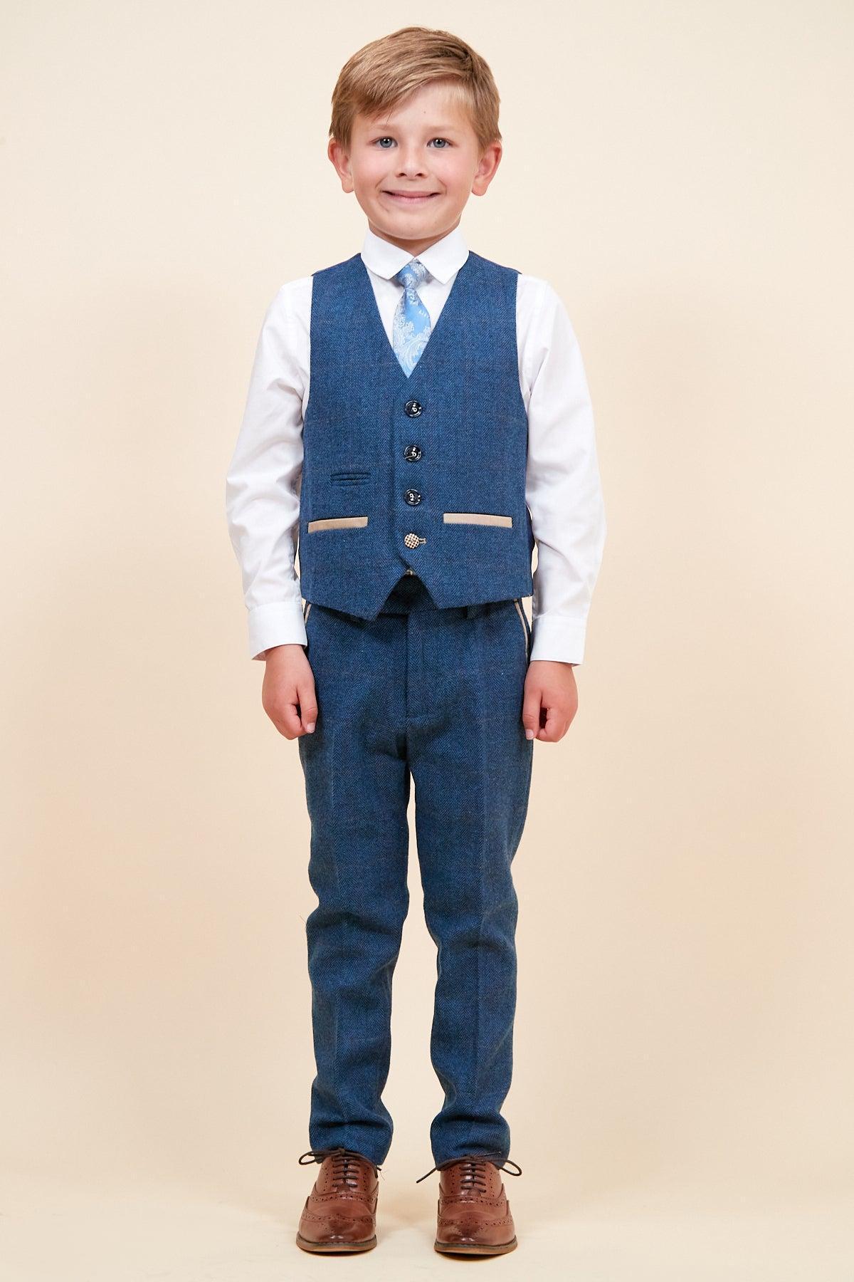 A young boy smiles in the marcdarcy DION Children's Blue Tweed Check Three Piece Suit, paired with a white shirt, blue patterned tie, and brown dress shoes, standing against a plain beige background.