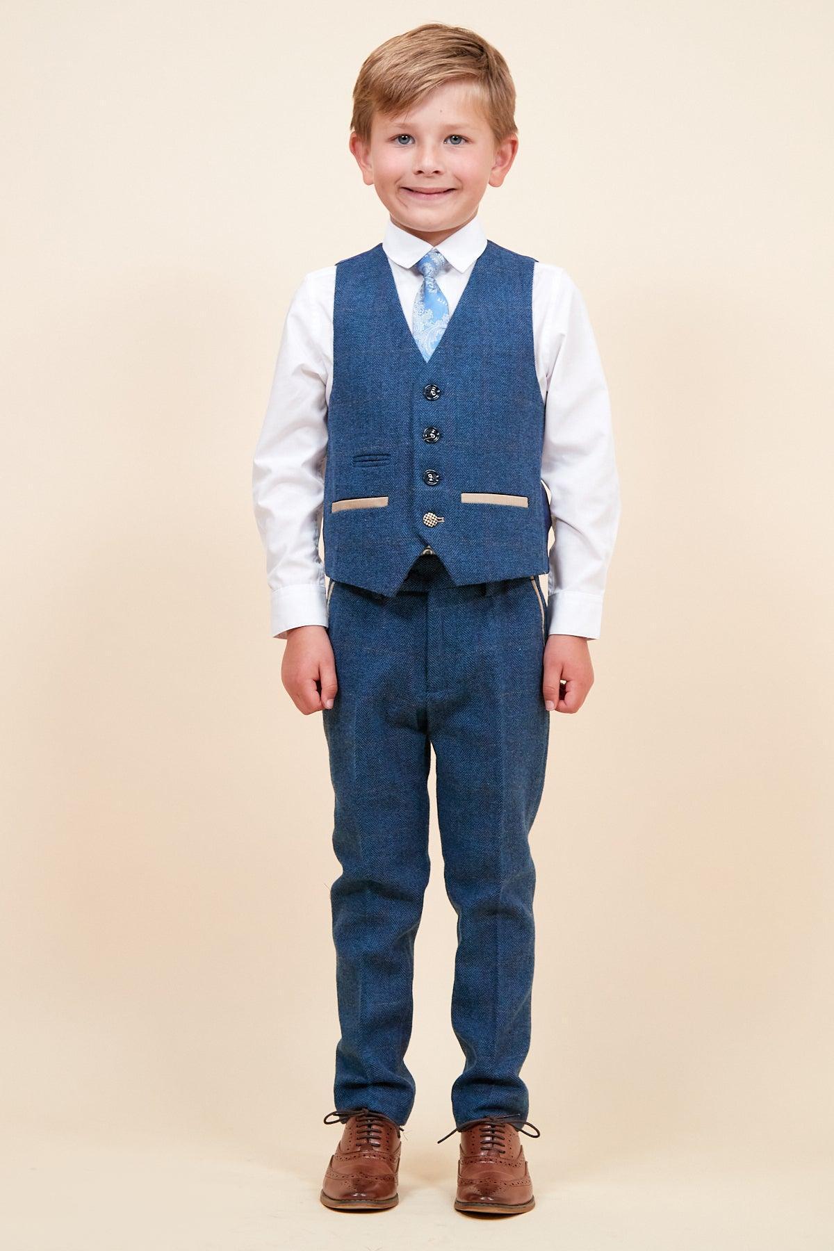 A young boy in a marcdarcy DION Children's Blue Tweed Check Three Piece Suit—paired with a white shirt, light blue patterned tie, and brown dress shoes—smiles at the camera.