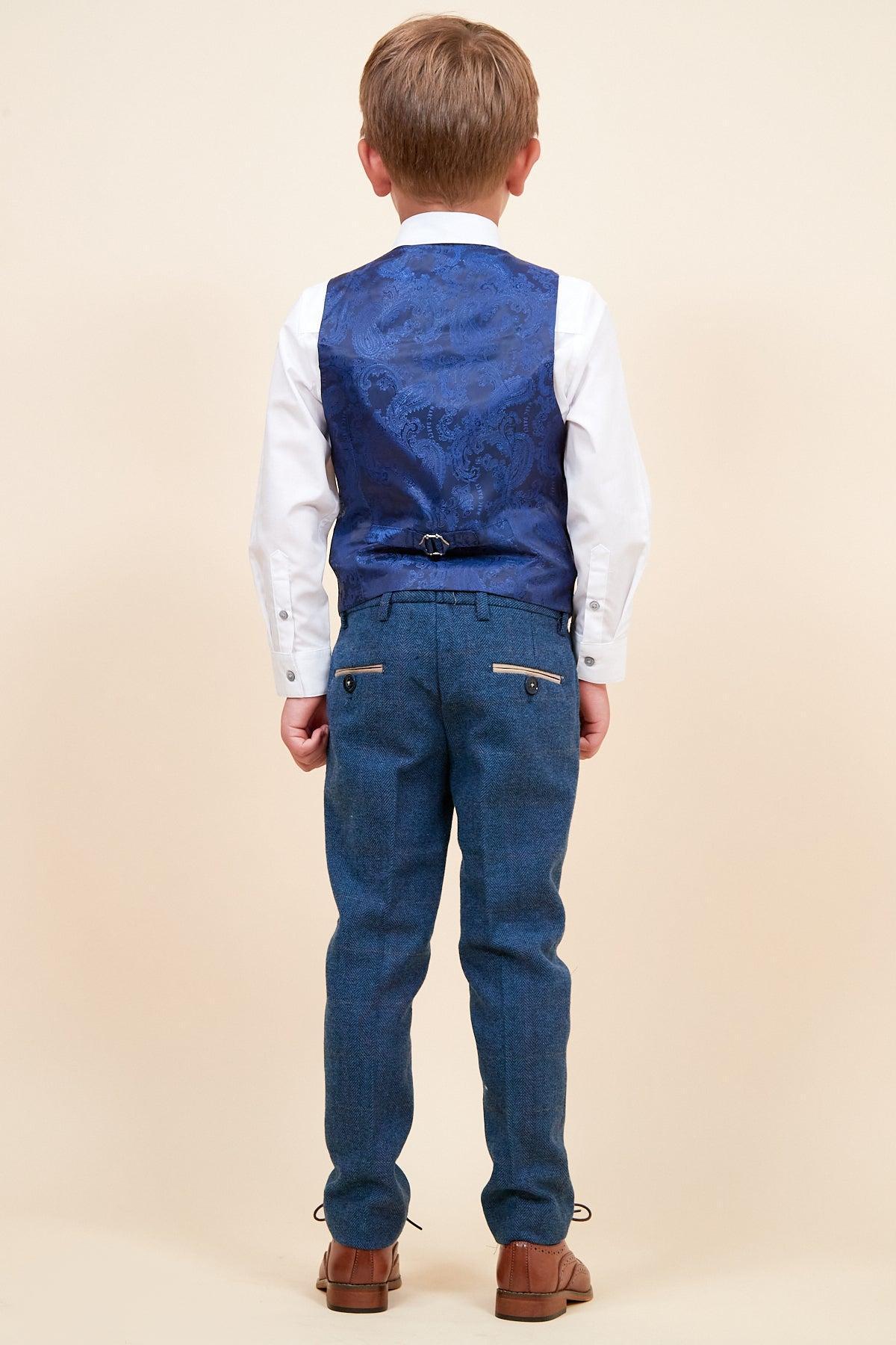 A young boy faces away, wearing the marcdarcy DION Childrens Blue Tweed Check Three Piece Suit with a blue patterned vest, blue dress pants, and brown shoes, set against a plain beige background.