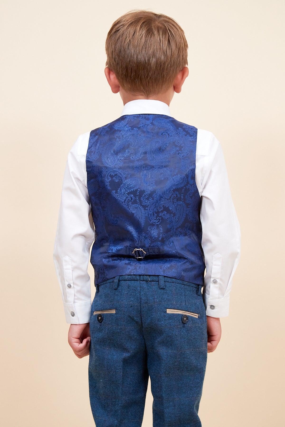 A young boy with light brown hair faces away, wearing the marcdarcy DION Children's Blue Tweed Check Three Piece Suit against a plain beige background—featuring a white shirt, blue patterned vest, and blue trousers with back pockets.