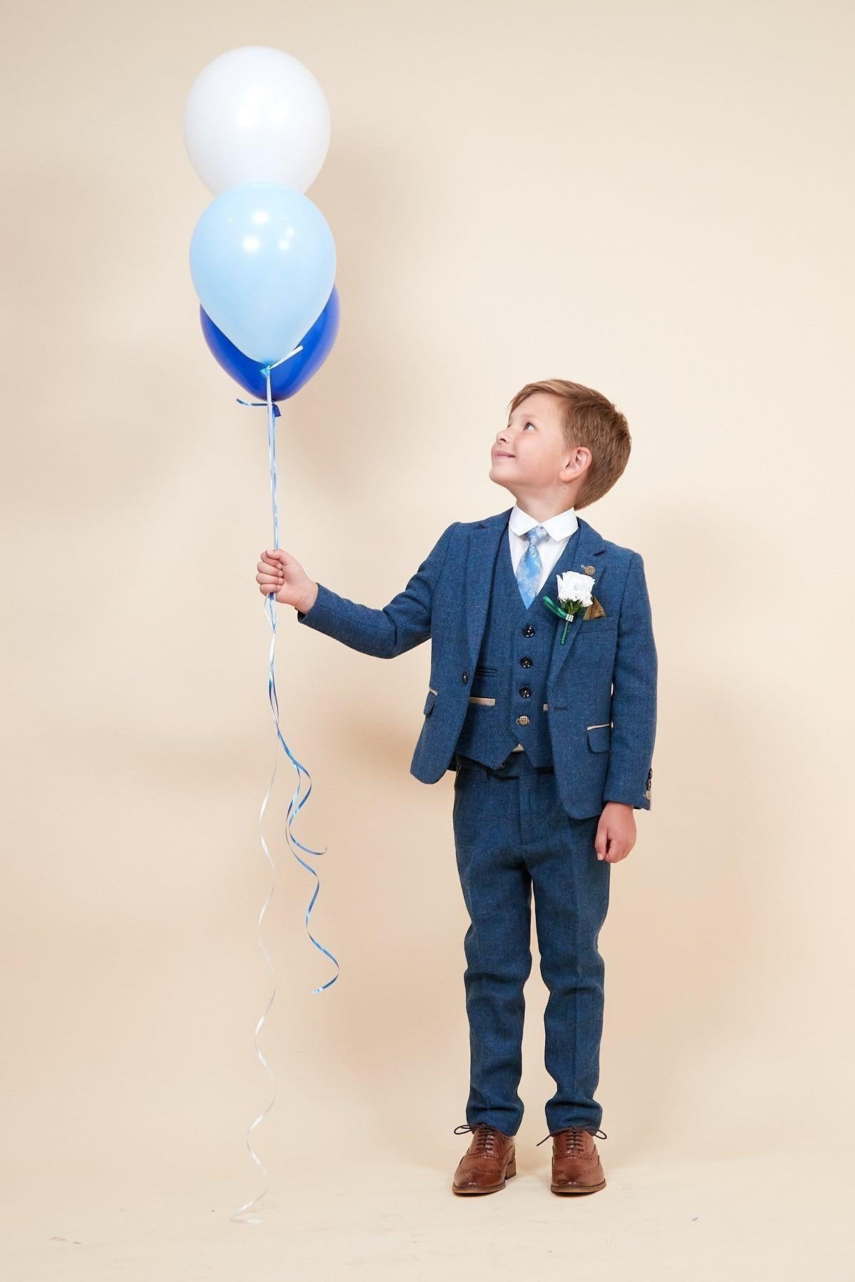 A young boy wearing the marcdarcy DION Children's Blue Tweed Check Three Piece Suit holds three blue and white balloons, looking up at them with a smile.