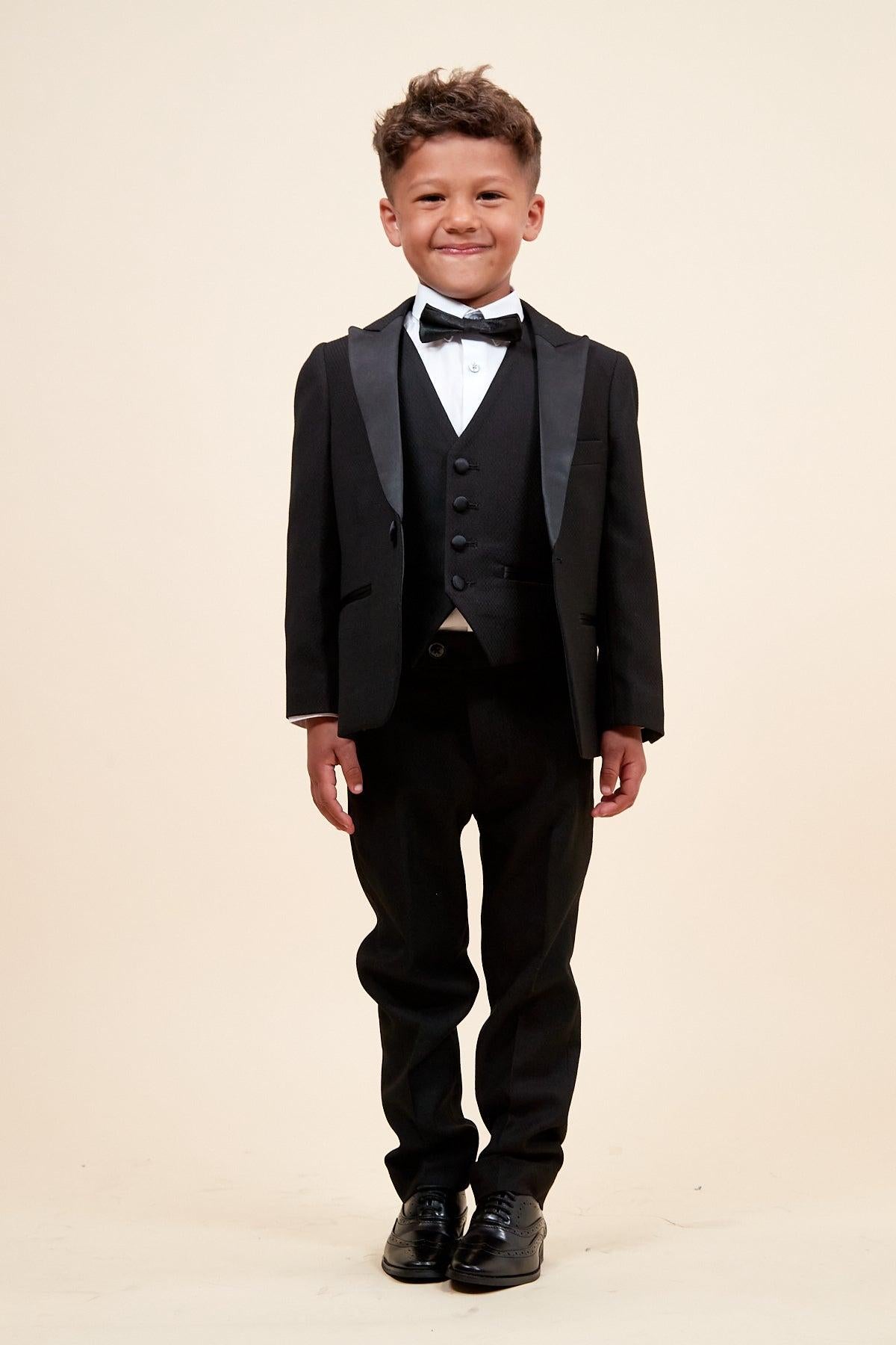 A young boy smiles in the marcdarcy Matching Father & Son | DALTON – Black Tux Three Piece Suit, featuring a bow tie, white shirt, vest, and black dress shoes, against a plain light beige background.
