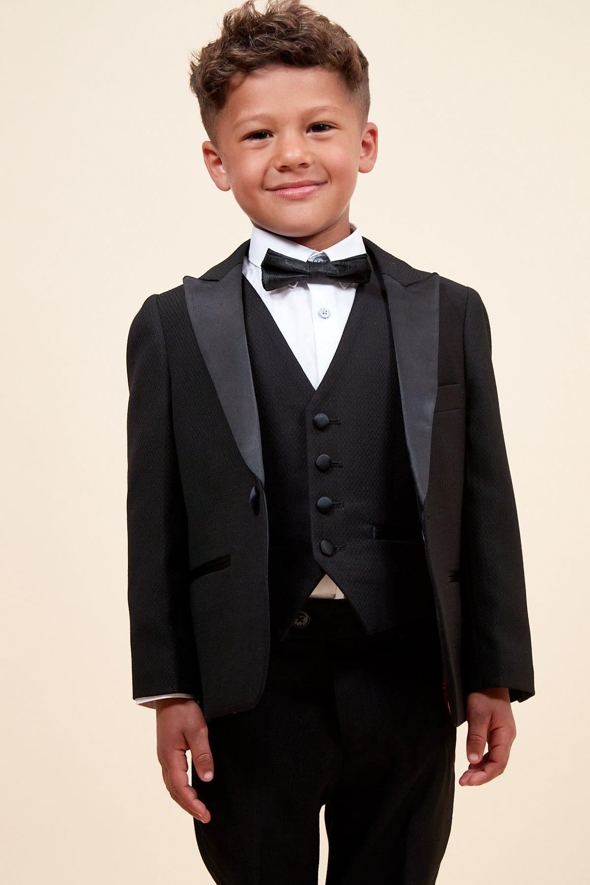 A young boy smiles in the marcdarcy Matching Father & Son | DALTON - Black Tux Three Piece Suit, featuring a bow tie and white dress shirt, set against a plain, light-colored background.