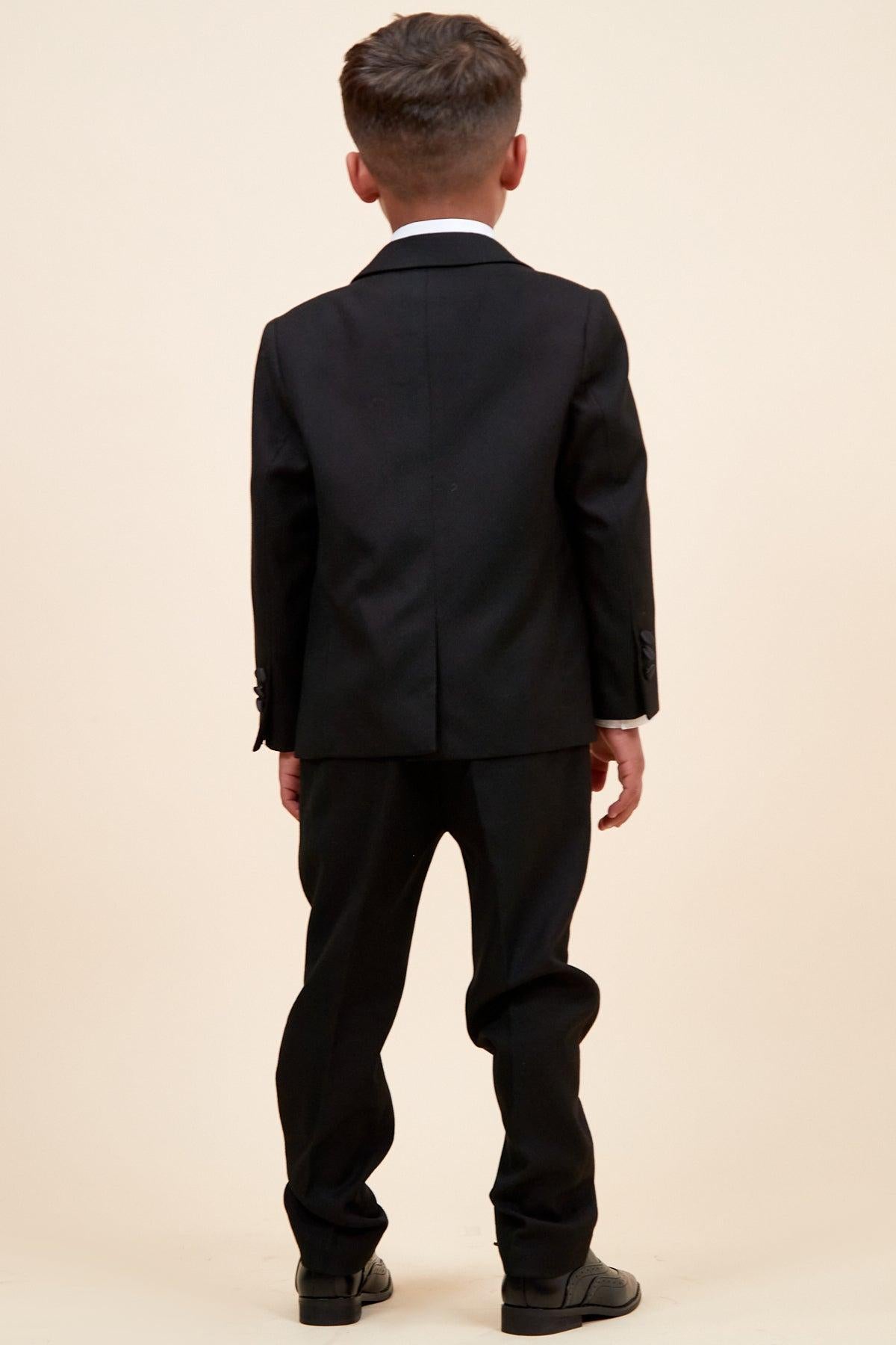A boy wears the marcdarcy Matching Father & Son | DALTON - Black Tux Three Piece Suit, featuring a blazer, trousers, and white shirt, paired with black shoes, photographed from behind against a cream background.