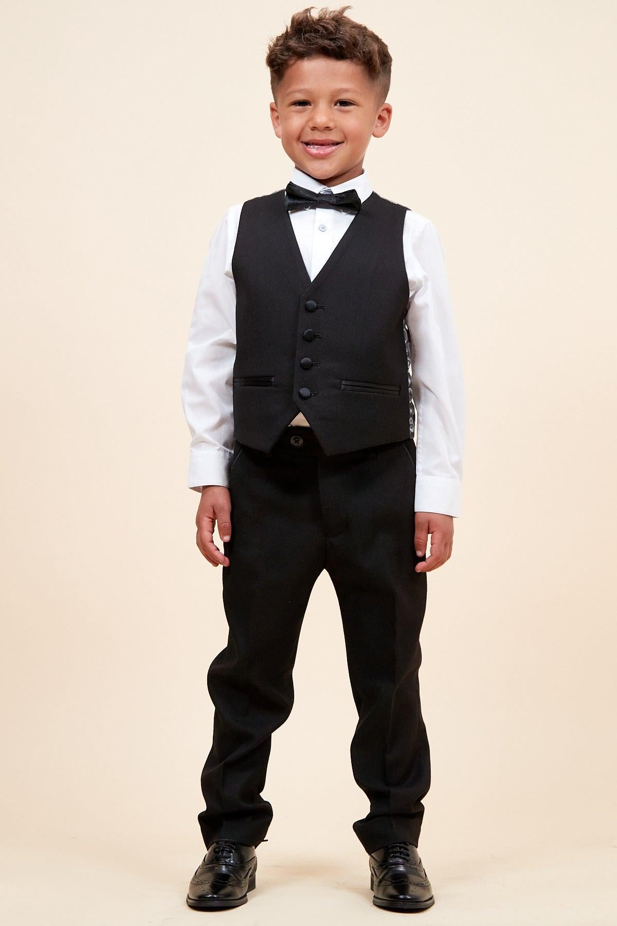 A young boy smiles in the marcdarcy DALTON Children's Black Tux Lapel Diamond Three Piece Suit, featuring a black vest and pants, white dress shirt, black bow tie, and dress shoes against a plain light background.