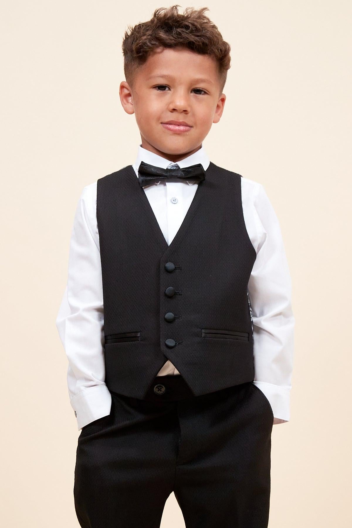 A young boy in the marcdarcy DALTON Children's Black Tux Lapel Diamond Three Piece Suit—featuring a crisp shirt, bow tie, vest, and pants—smiles confidently with his hands in his pockets against a plain background.