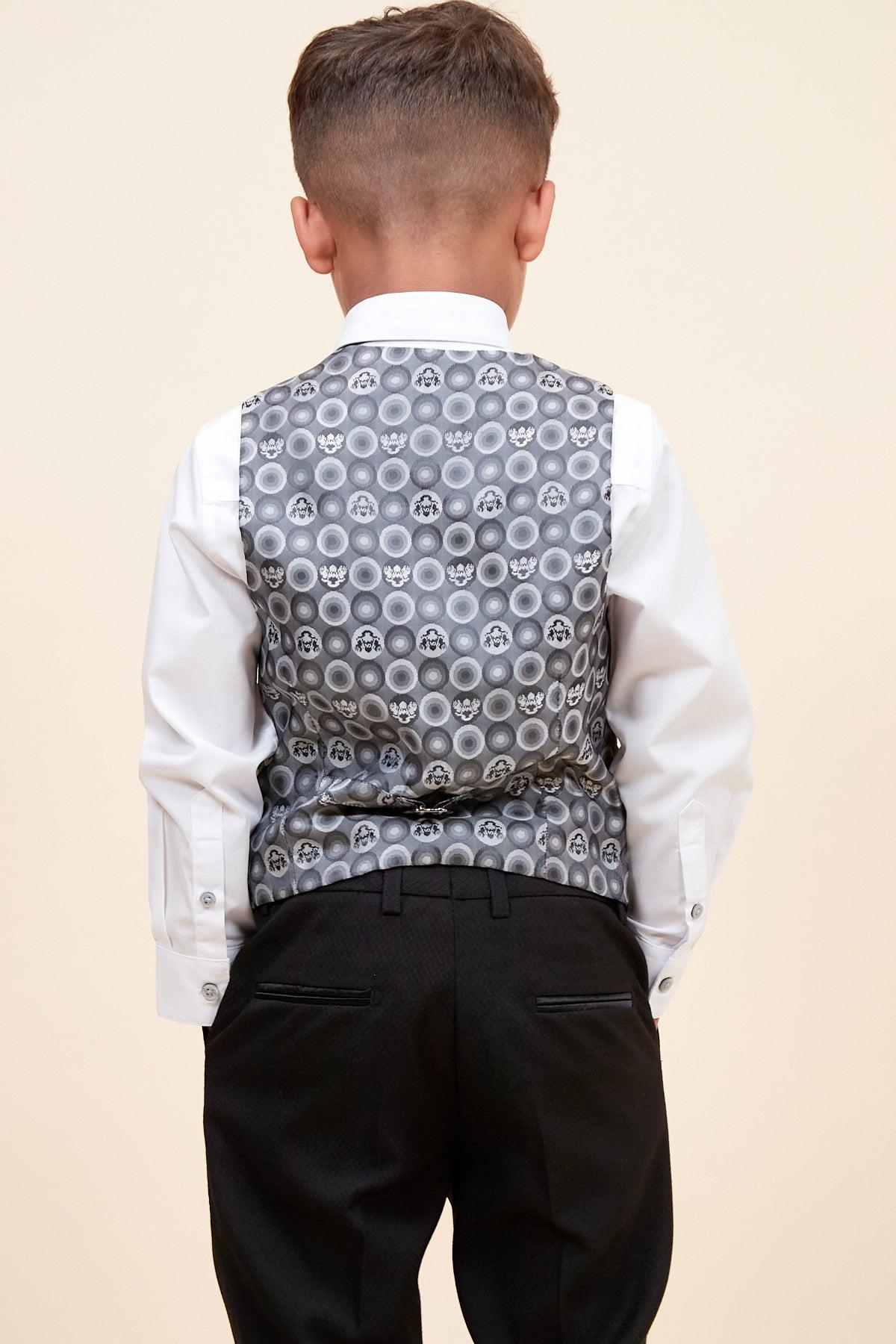 Back view of a boy wearing the marcdarcy DALTON Children's Black Tux Lapel Diamond Three Piece Suit: white shirt, grey patterned vest with circles and faces, and black dress pants.