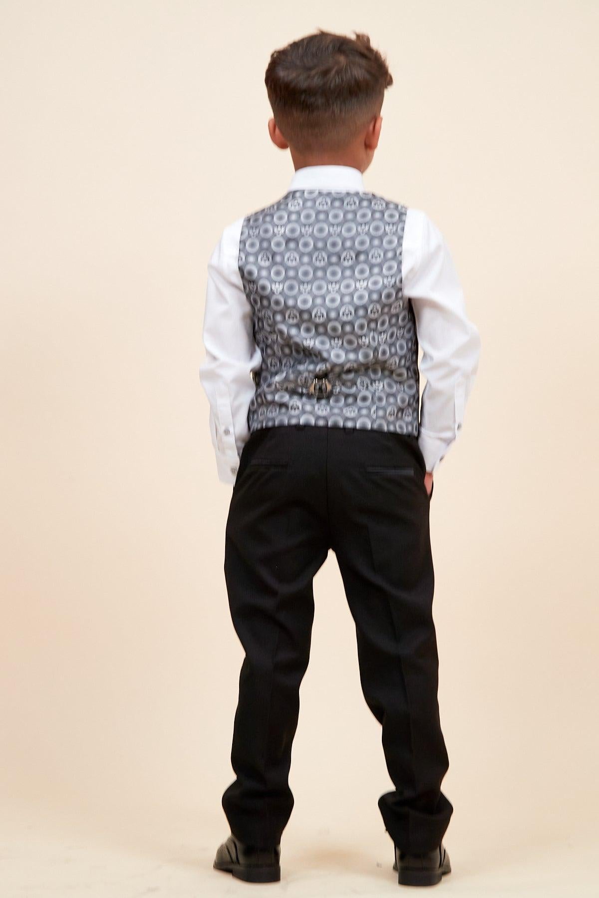 A young boy stands facing away, wearing the marcdarcy DALTON - Children's Black Tux Lapel Diamond Three Piece Suit, featuring a white shirt, silver patterned vest, and black pants, set against a plain beige background.