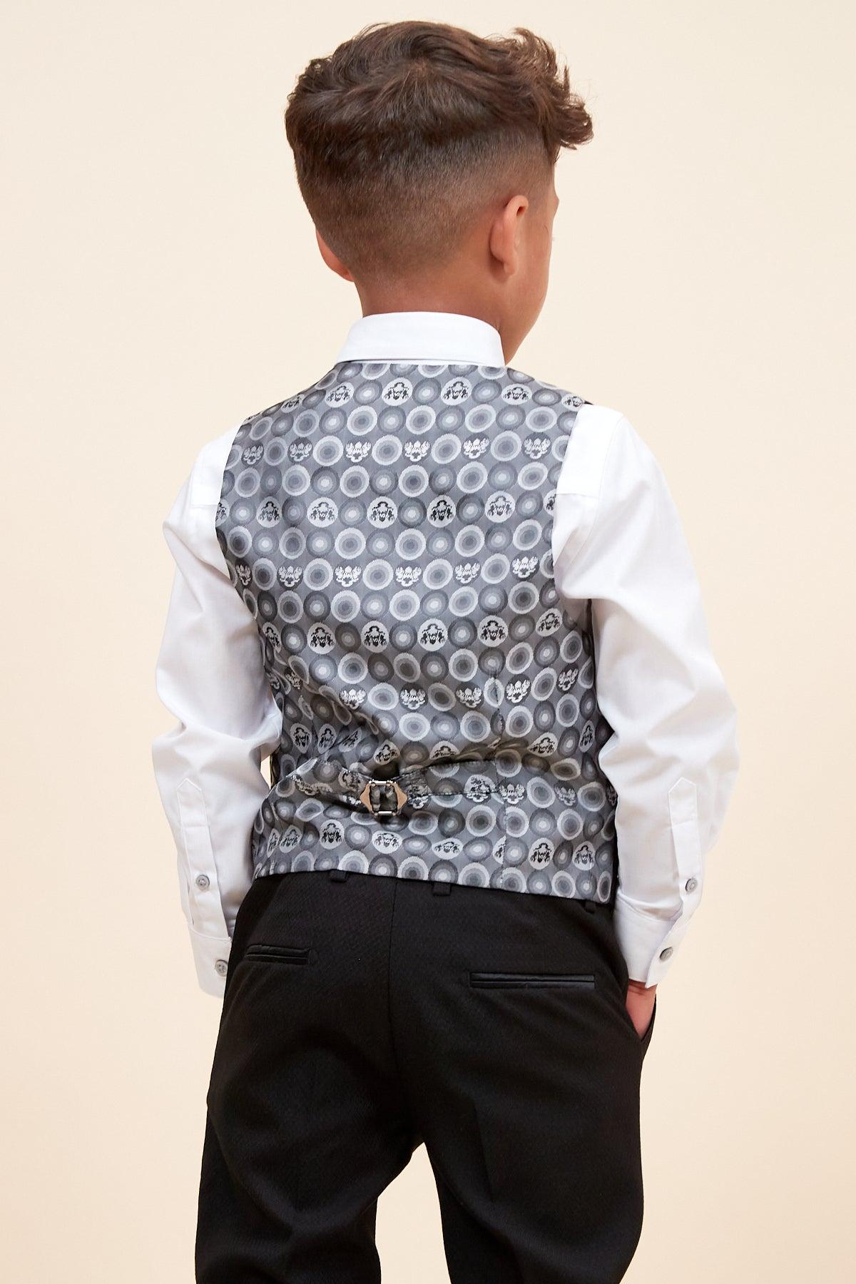 A young boy stands with his back to the camera, hands in pockets, wearing the DALTON - Children's Black Tux Lapel Diamond Three Piece Suit by marcdarcy: a classic white shirt, black trousers, and a silver-gray patterned vest.