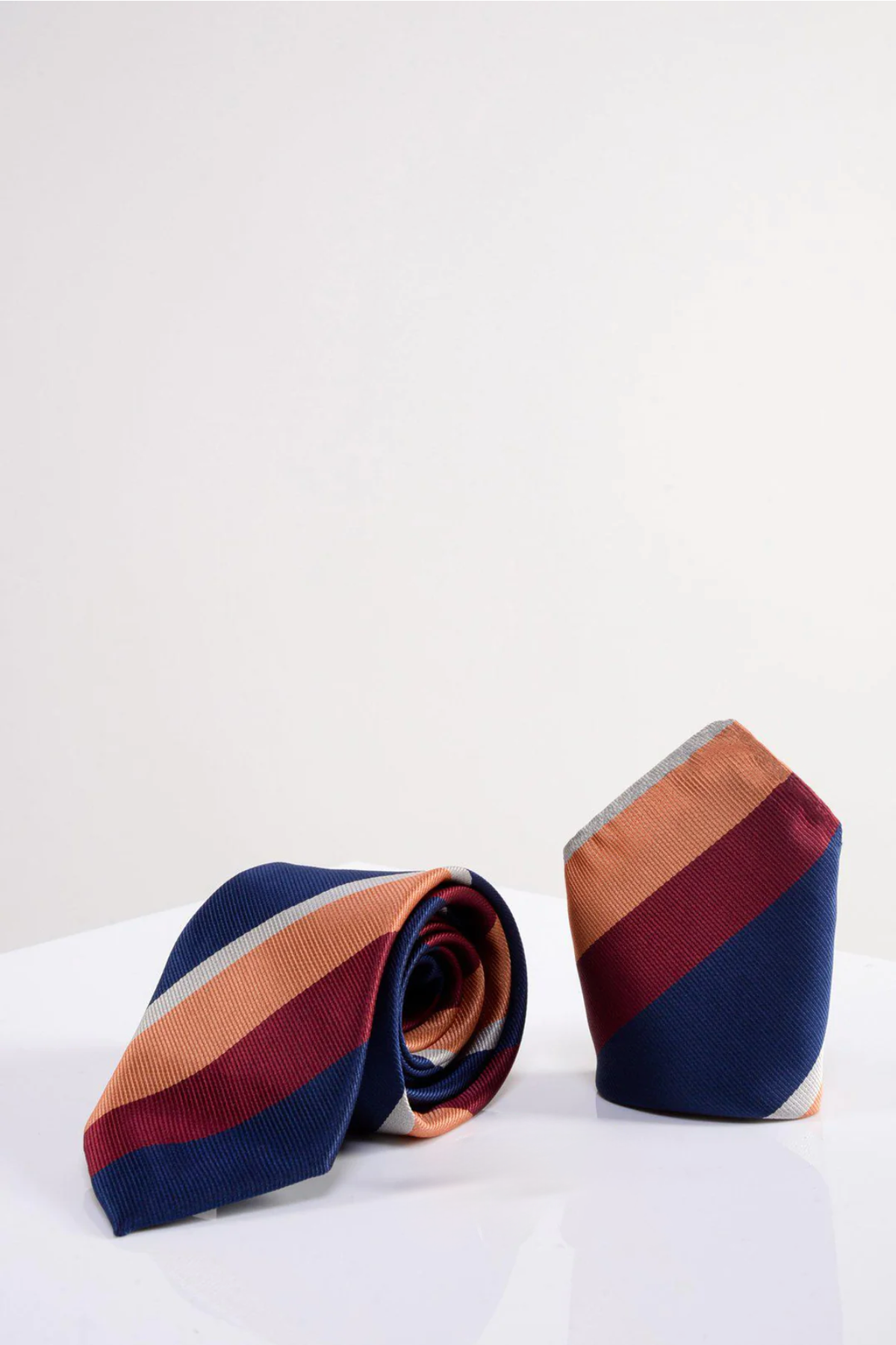 The Marc Darcy BRUNO Multicolour Stripe Print Tie and Pocket Square Set features two ties with diagonal navy, orange, and burgundy stripes on a white surface—one rolled, one upright—adding bold flair to any formal outfit.