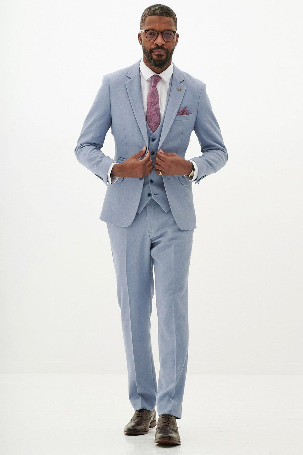 A man in the Marc Darcy HM5 Sky Blue Three Piece Suit with a white shirt, purple patterned tie, and brown shoes stands against a plain background. Short-haired with a beard and glasses, he showcases this modern wedding suit's elegance.