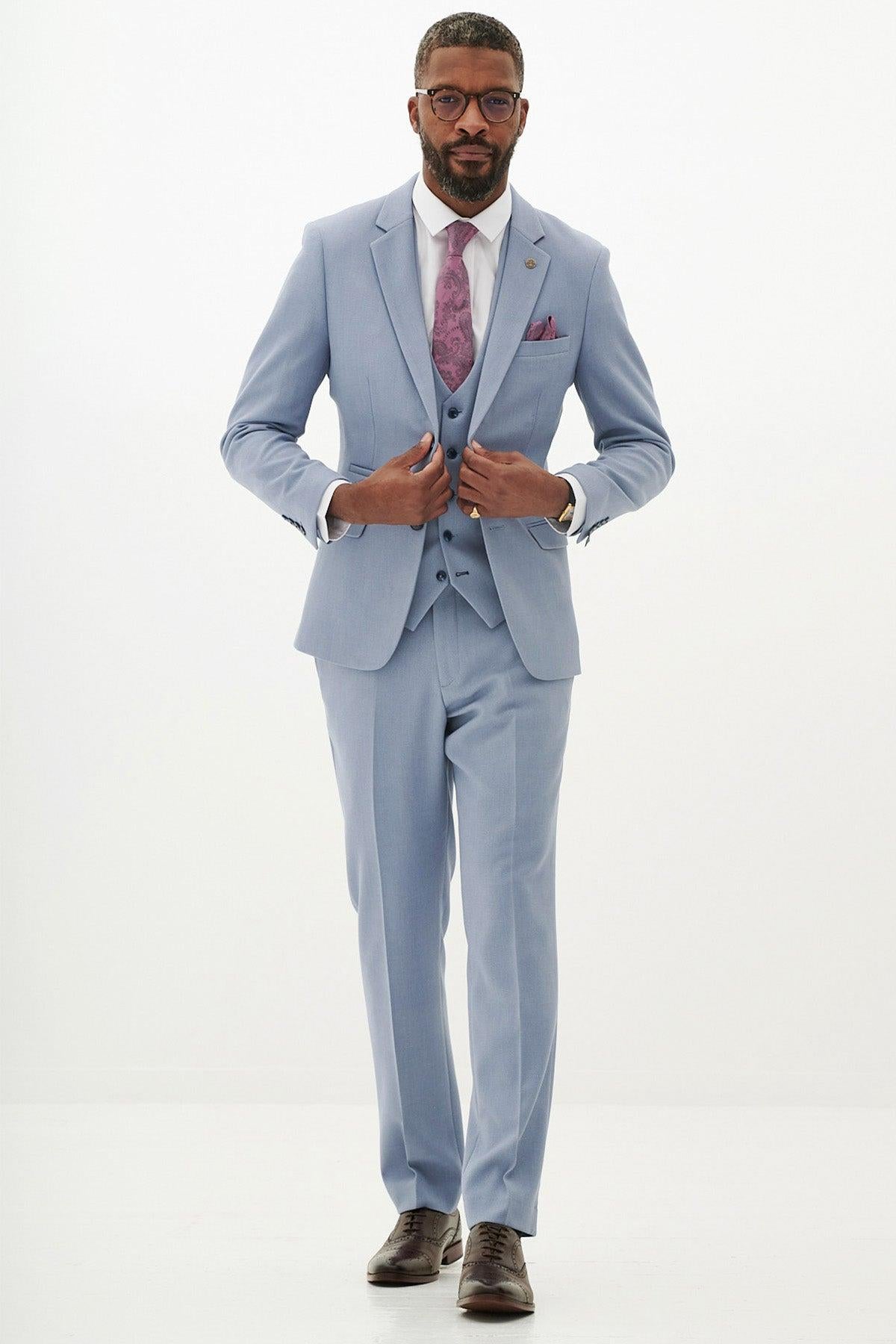 A man in the Marc Darcy HM5 Sky Blue Three Piece Suit with a white shirt, purple patterned tie, and brown shoes stands against a plain background. Short-haired with a beard and glasses, he showcases this modern wedding suit's elegance.
