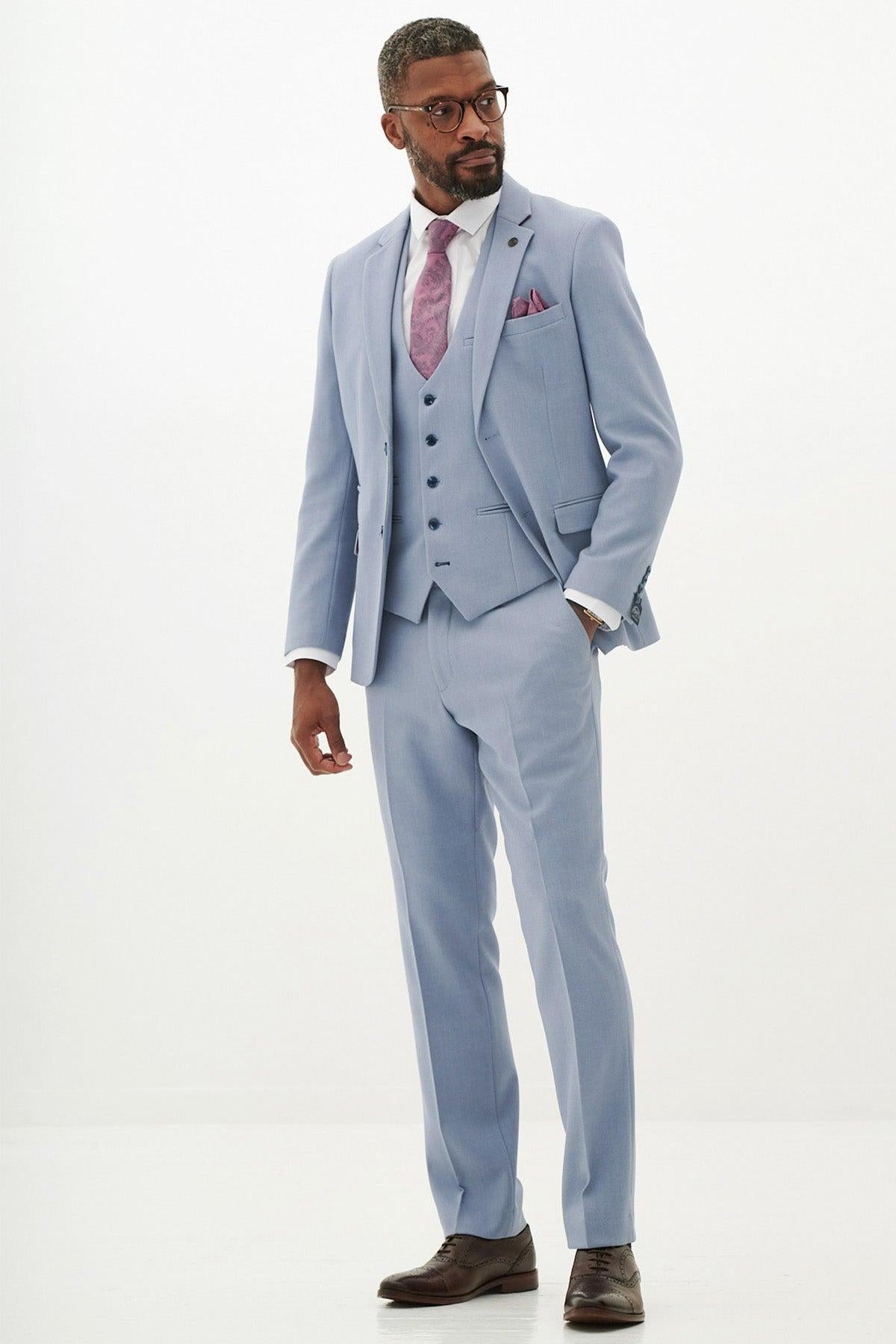 A man wearing the Marc Darcy HM5 - Sky Blue Three Piece Suit with a white shirt, mauve tie, and pocket square stands against a white background. He has short hair, glasses, and brown dress shoes.
