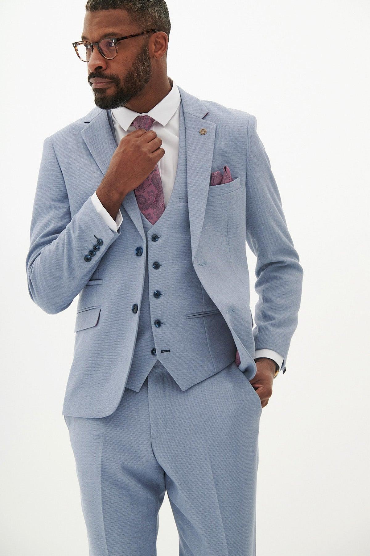 A man with glasses and a beard adjusts his tie, wearing the Marc Darcy HM5 - Sky Blue Three Piece Suit paired with a patterned pink tie and pocket square—a stylish men's wedding suit choice.