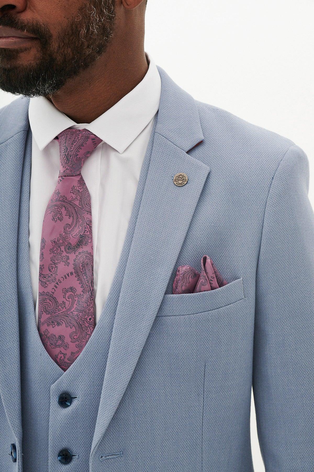 A man in the Marc Darcy HM5 - Sky Blue Three Piece Suit with a white shirt, pink paisley tie, pocket square, and lapel pin—an ideal wedding outfit for a stylish celebration.