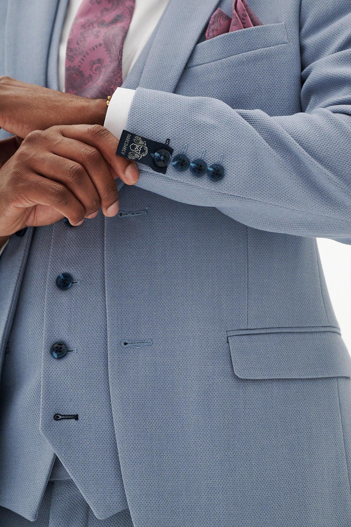 A man wearing the Marc Darcy HM5 - Sky Blue Three Piece Suit adjusts his sleeve, showing the label. He pairs it with a matching vest, pink patterned tie, and pink pocket square. The photo highlights the suit's details and his hands.