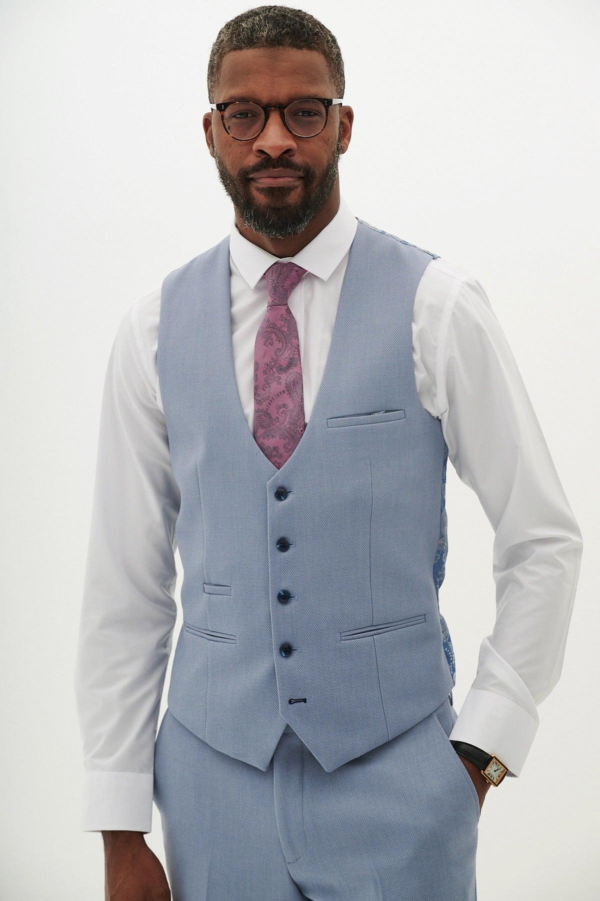 A man wearing glasses, the marcdarcy HM5 - Sky Blue Single Breasted Waistcoat, matching trousers, a white dress shirt, and a mauve patterned tie stands with one hand in his pocket against a plain white background.