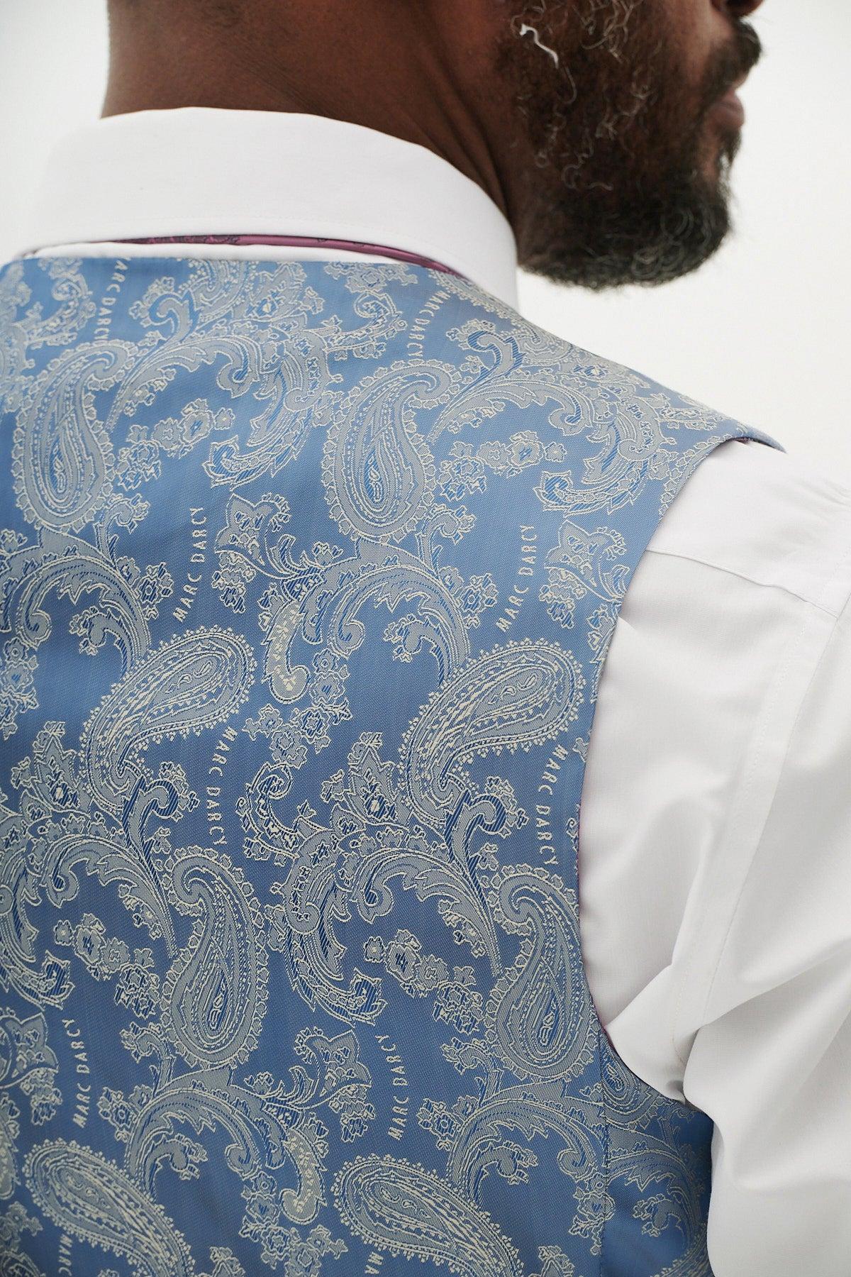A man wears the marcdarcy HM5 Sky Blue Single Breasted Waistcoat with a paisley pattern over a white shirt, featuring a subtle brand logo—an elegant choice for weddings and special occasions.