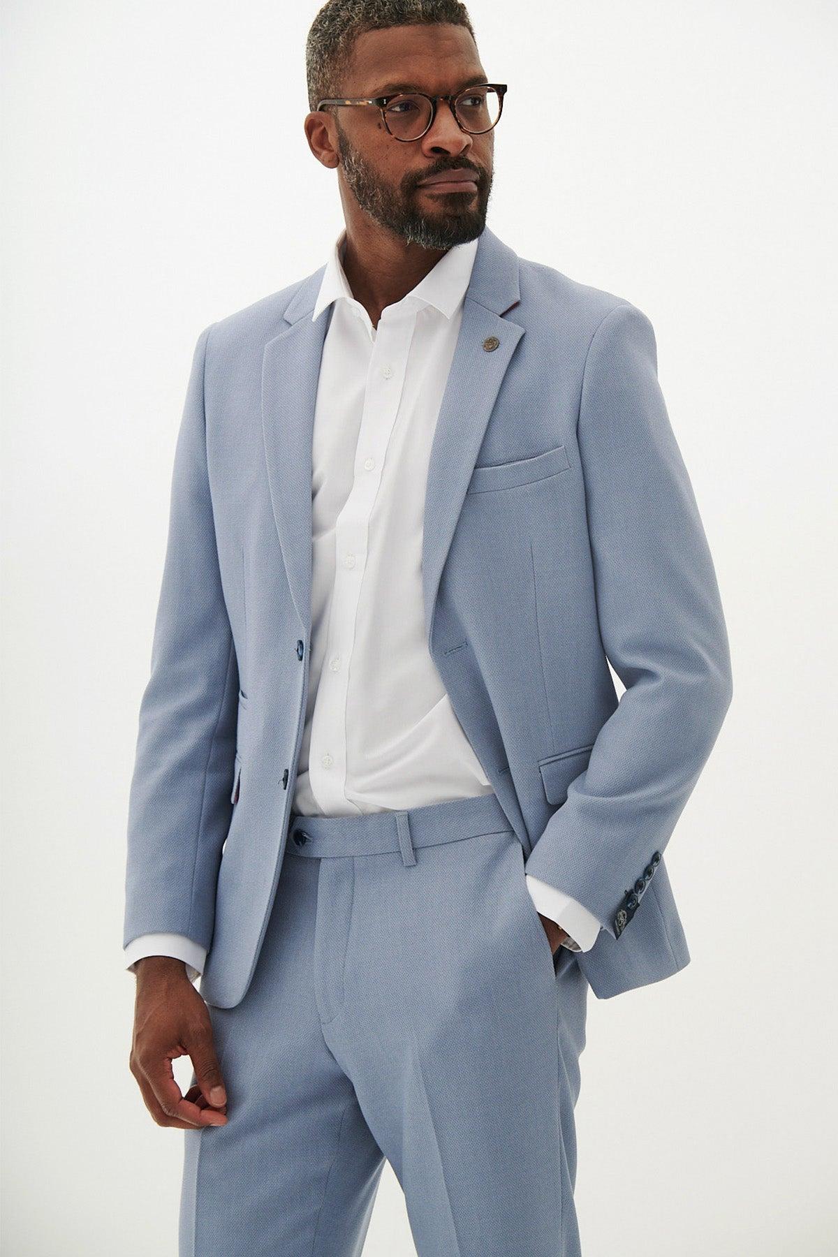 A man models the Marc Darcy HM5 - Sky Tailored Two Piece Suit with a white shirt and glasses, standing against a plain white background and looking slightly to the side with one hand in his pocket.