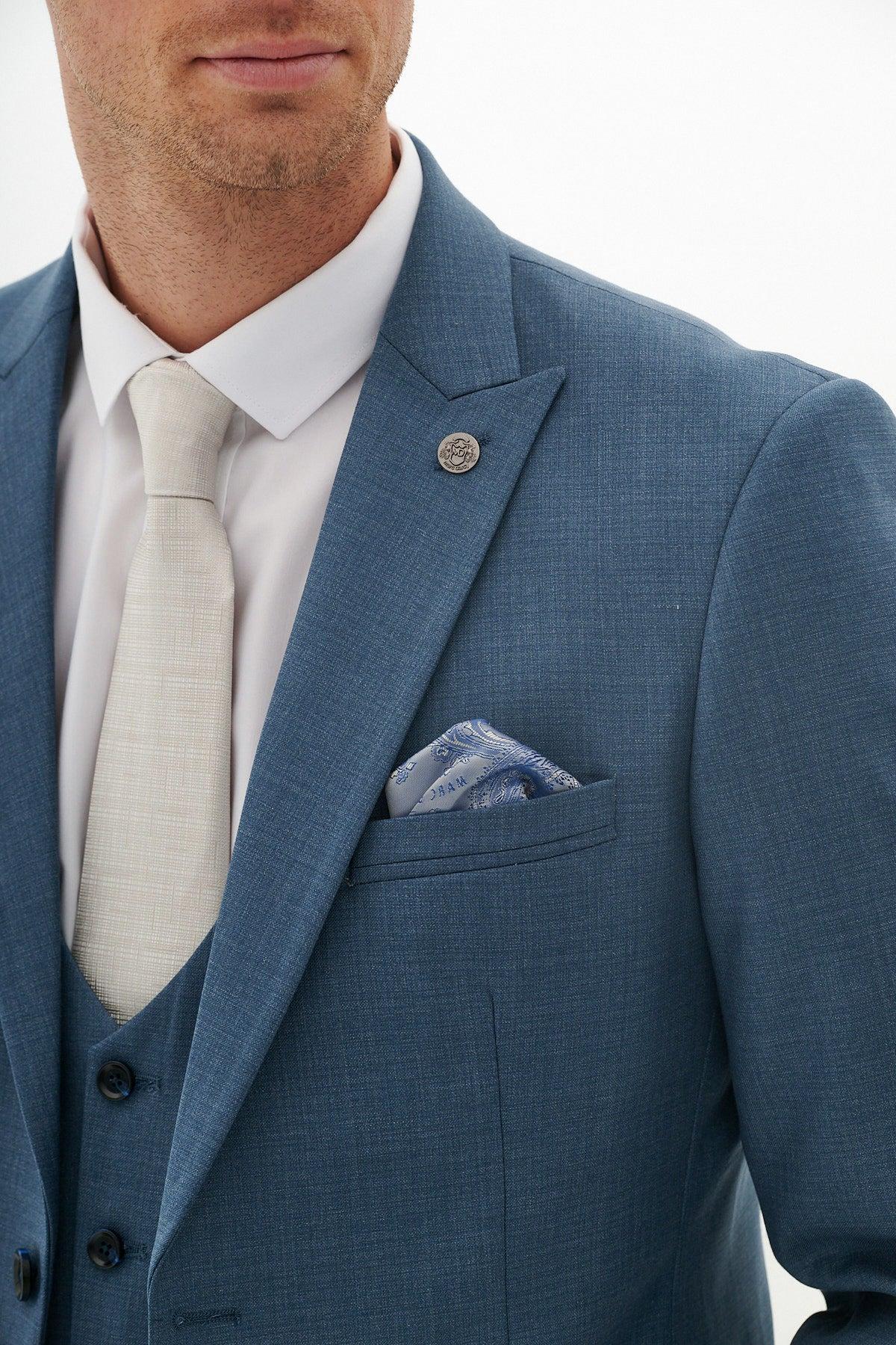 A man wears the Marc Darcy FINLEY teal blue three-piece suit, styled with a white shirt, cream tie, blue pocket square, and lapel pin—ideal for a formal wedding look.