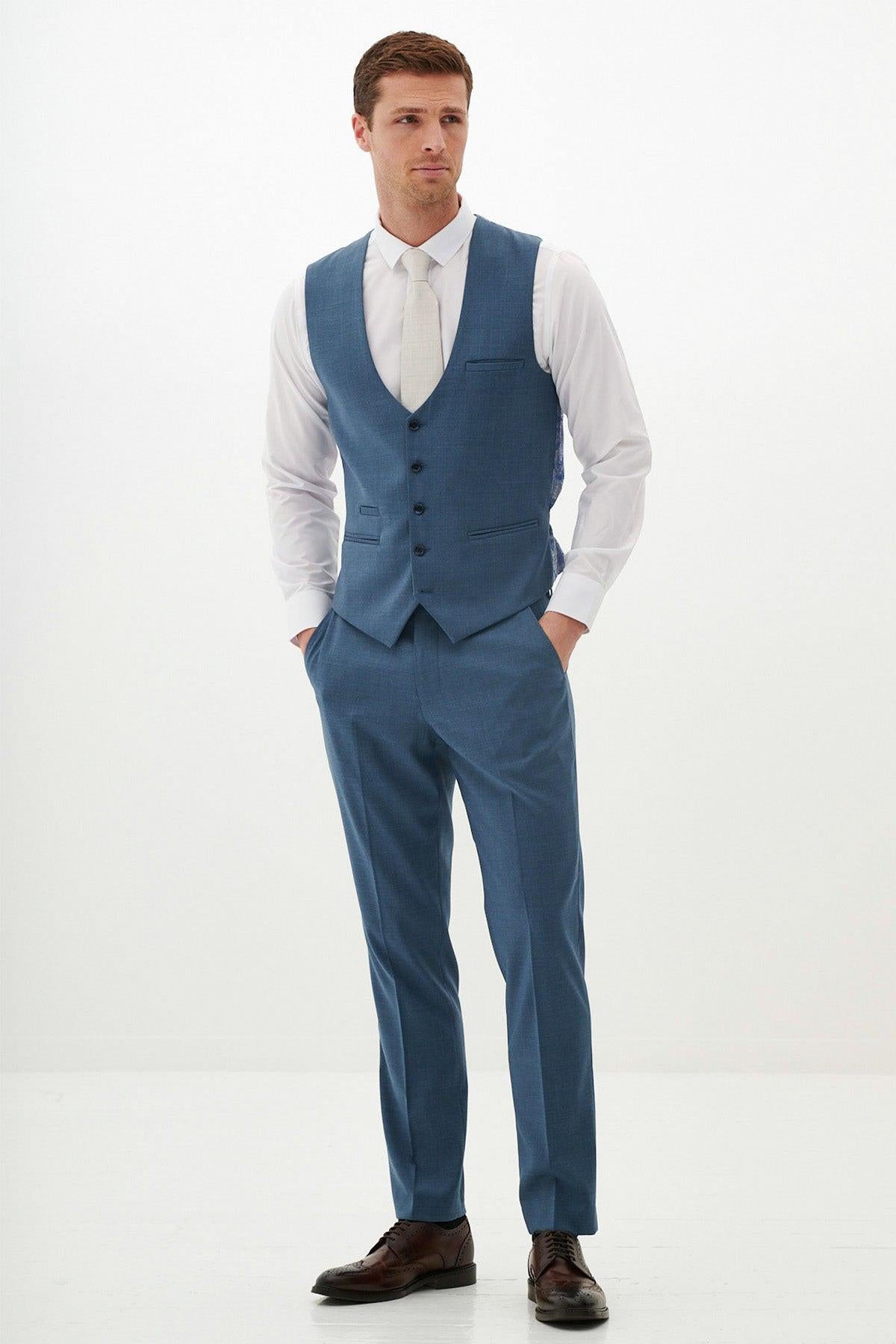 A man stands with his hands in his pockets, wearing the Marc Darcy FINLEY Teal Blue Single Breasted Waistcoat over a white dress shirt, paired with a light gray tie and brown dress shoes, against a plain white background.