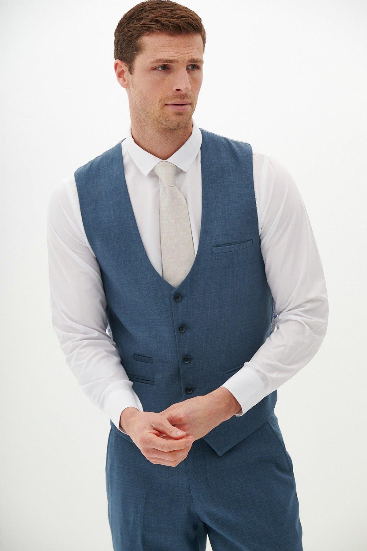 A man wears the FINLEY Teal Blue Single Breasted Waistcoat by Marc Darcy with a white shirt and cream tie, standing against a white background—a perfect summer formalwear look.