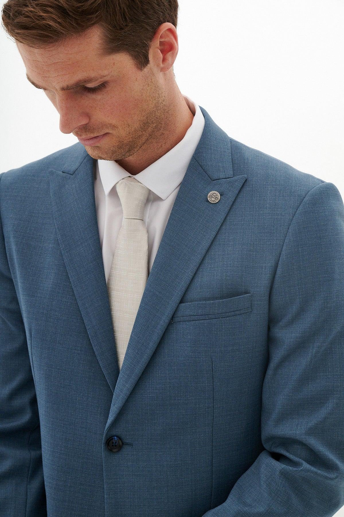 A man wearing the Marc Darcy FINLEY - Teal Blue Tailored Blazer, paired with a white shirt and beige tie, looks down. A lapel pin accents his suit jacket.