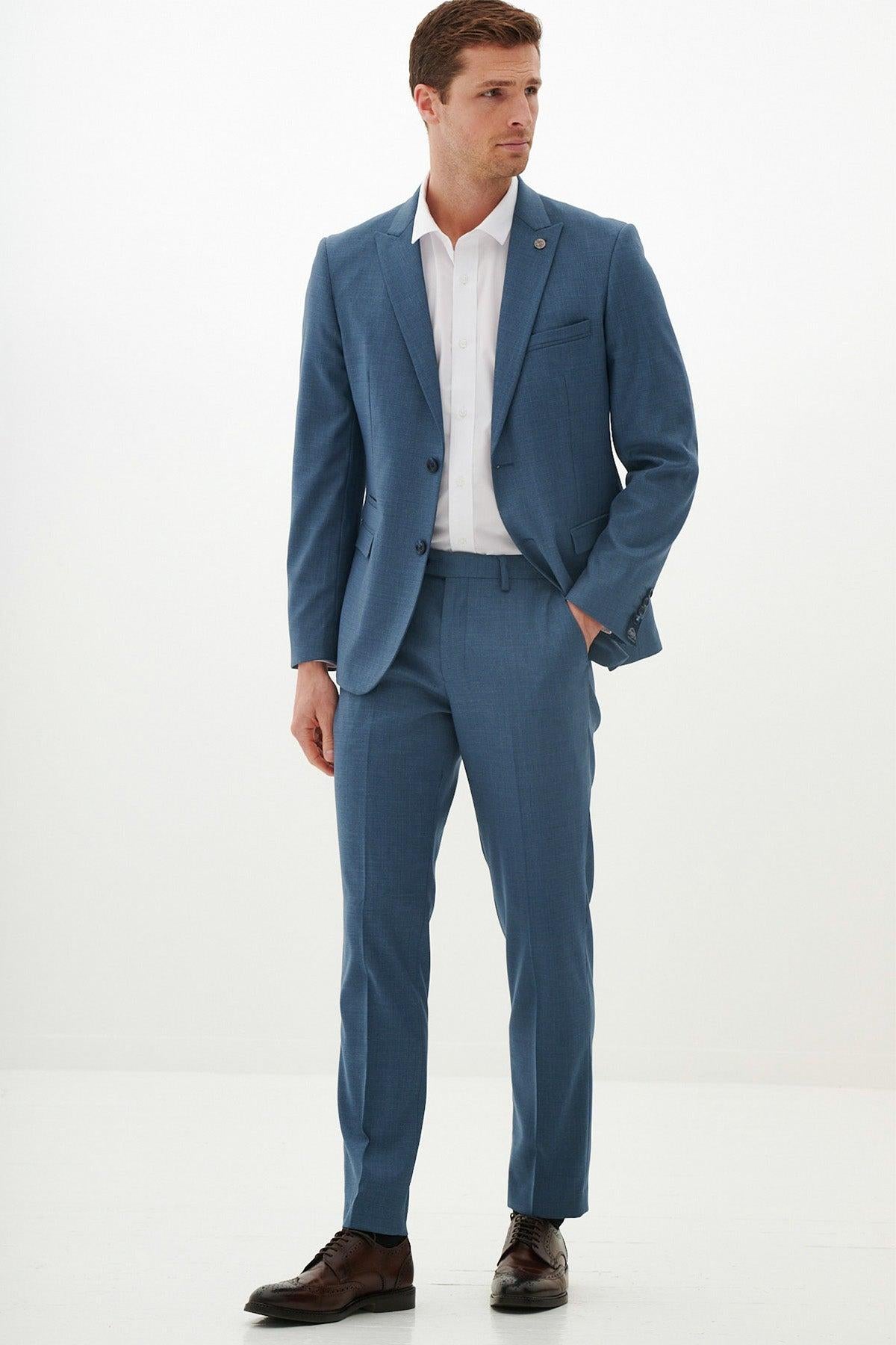A man stands against a plain white background wearing the Marc Darcy FINLEY Teal Blue Two Piece Suit with a white shirt and brown dress shoes, looking to the side with one hand in his pocket—a sharp summer wedding outfit.