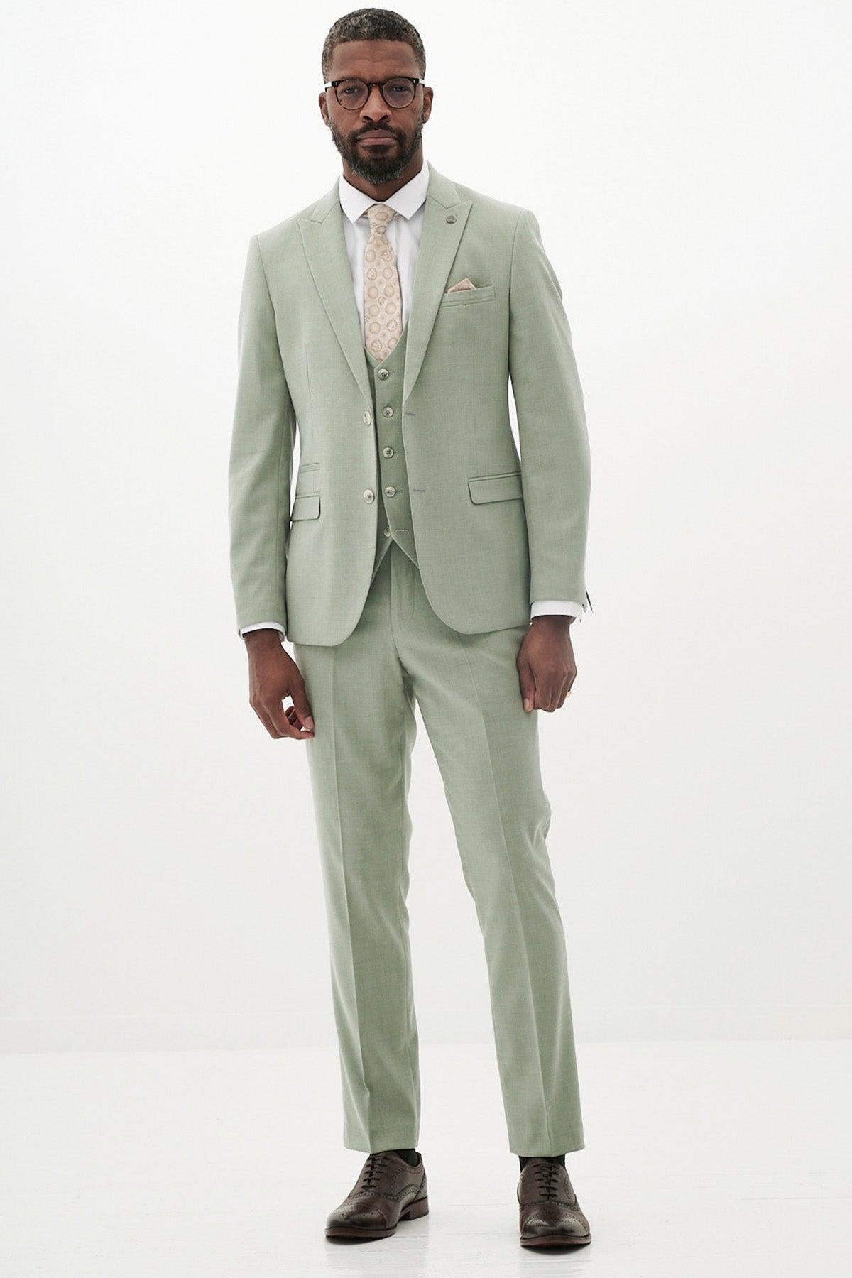 A Black man wearing the Marc Darcy FINLEY Moss Green Three Piece Suit, a white shirt, patterned tie, and brown shoes stands against a white background.