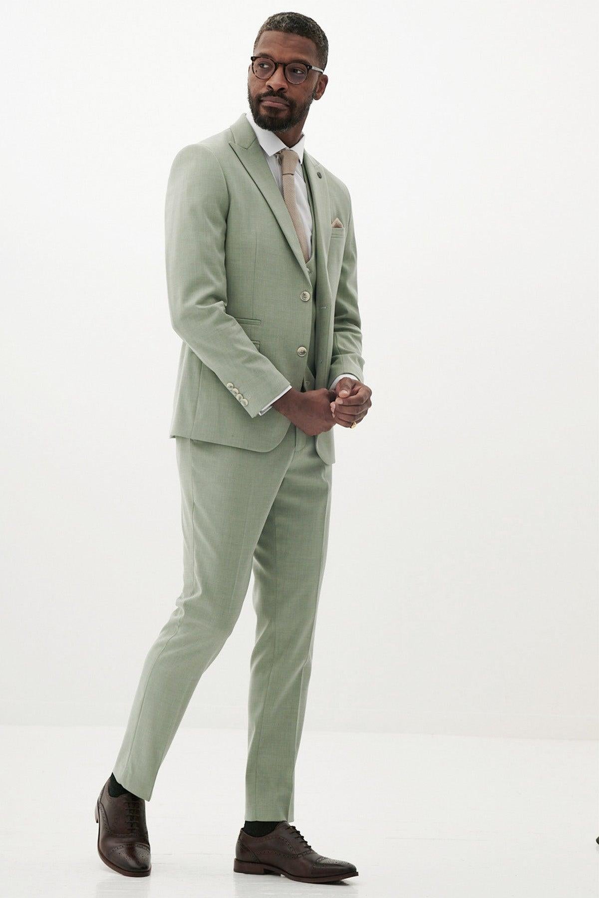 A man in the Marc Darcy FINLEY Moss Green Three Piece Suit—paired with a light dress shirt, beige tie, and brown shoes—stands against a plain white background, looking slightly to his left with his hands clasped.