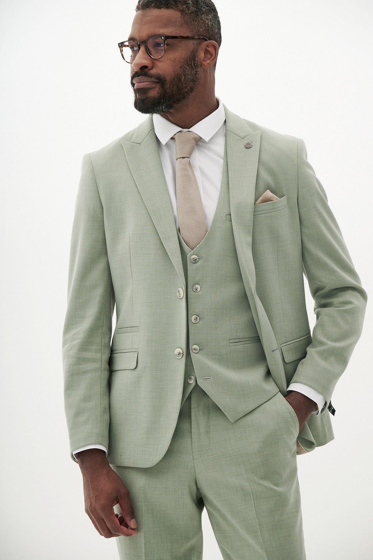 A man in a Marc Darcy FINLEY Moss Green Three Piece Suit, styled with a white shirt, beige tie, and glasses, stands against a plain white background—an ideal outfit for summer weddings.