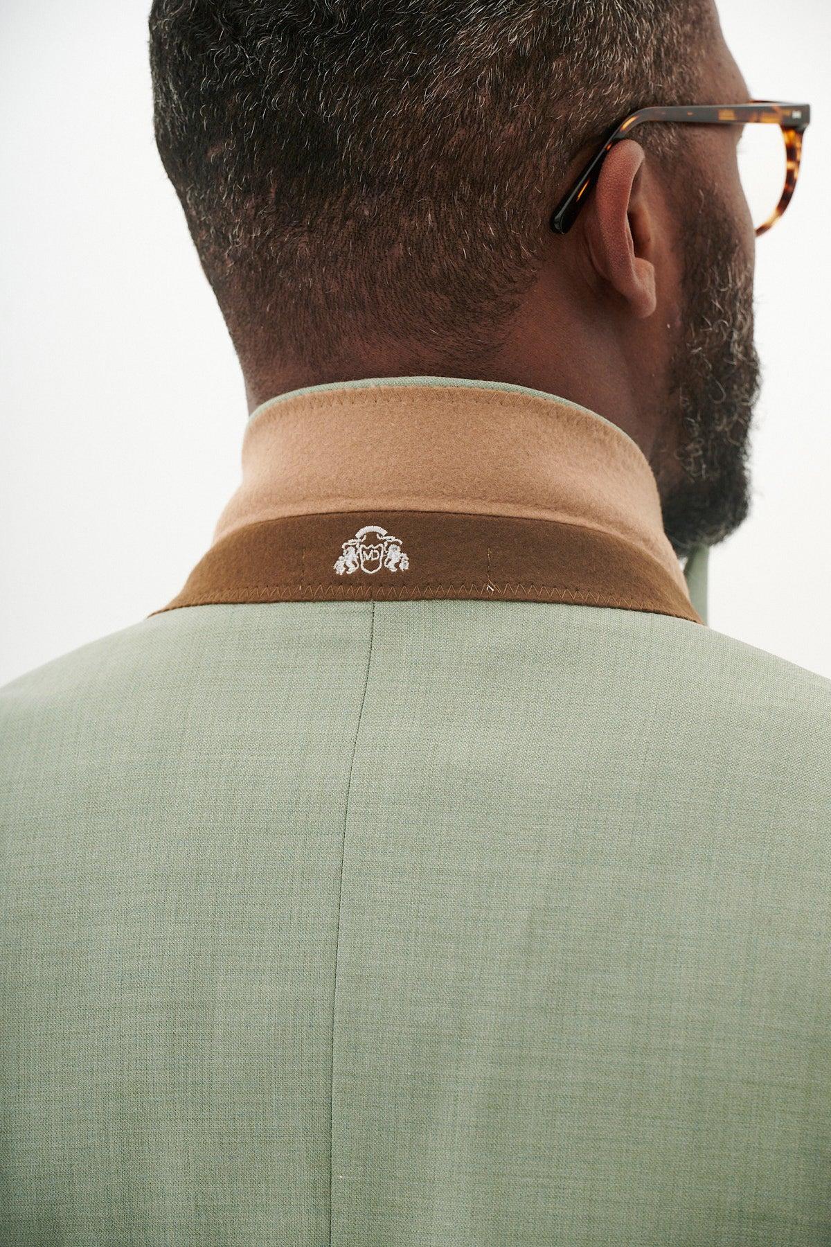 Close-up from behind of a Black man wearing the FINLEY - Moss Green Two Piece Suit by Marc Darcy, featuring a brown and tan collar with embroidered crest—lightweight and ideal for summer weddings.