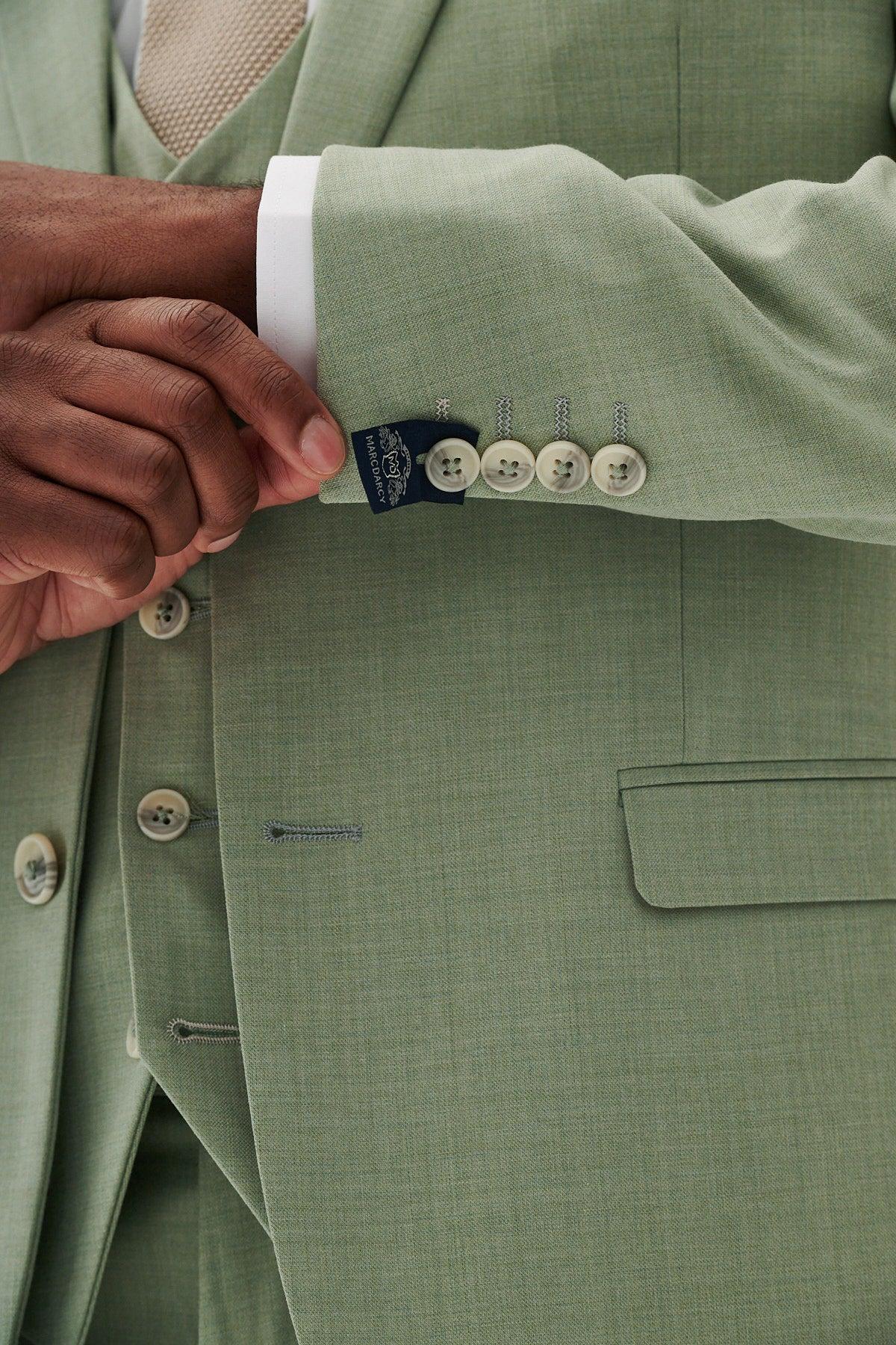 Wearing the Marc Darcy FINLEY - Moss Green Two Piece Suit, a person adjusts their left sleeve to reveal a label and light buttons. Paired with a matching vest, white shirt, and beige tie, its perfect for summer weddings.