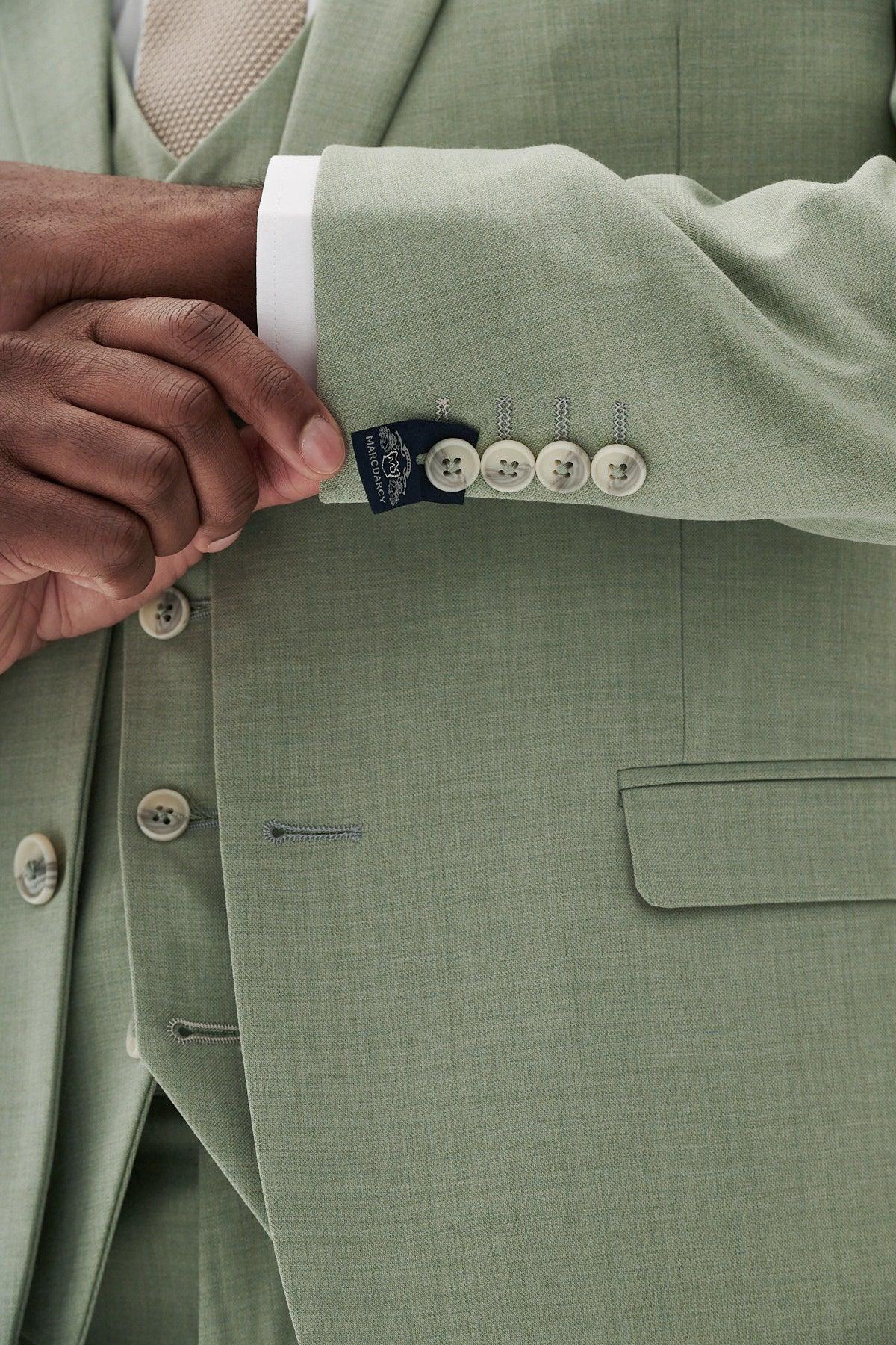 A close-up shows someone adjusting their white shirt cuff in the Marc Darcy FINLEY - Moss Green Three Piece Suit, featuring light buttons, a visible sleeve tag, and a buttoned vest underneath.