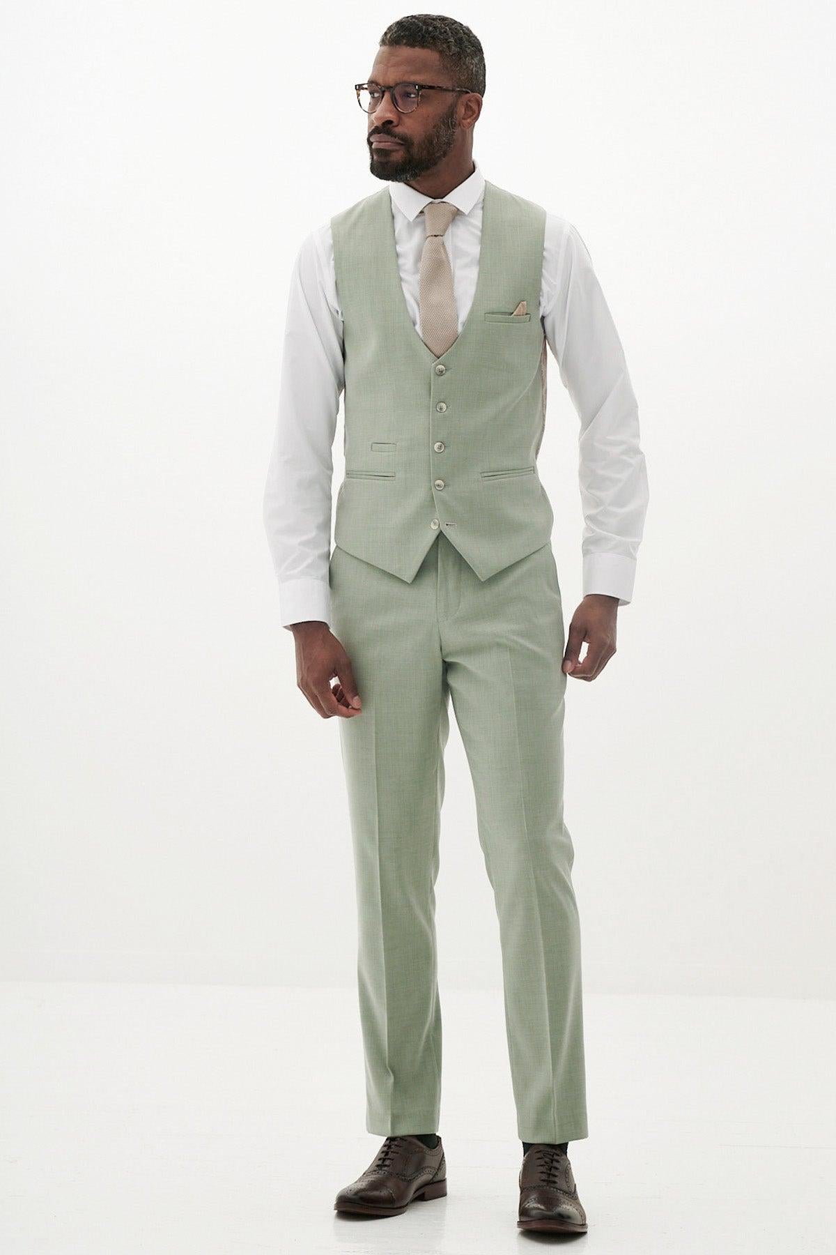 A man in glasses and a light sage green three-piece suit featuring the Marc Darcy FINLEY - Moss Green Single Breasted Waistcoat, white shirt, beige tie, and brown shoes stands against a white background—ideal summer formalwear.
