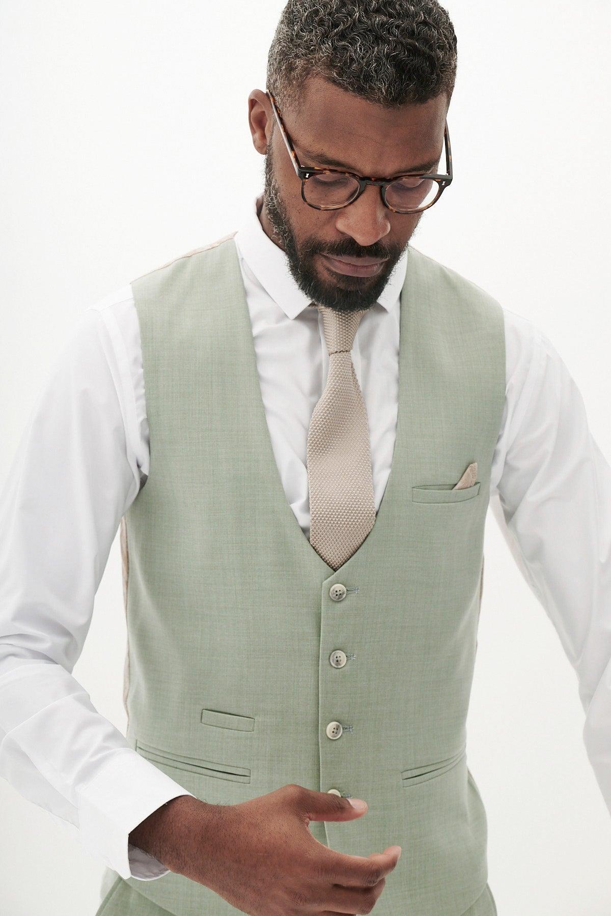 A man in glasses and a white shirt wears the Marc Darcy FINLEY - Moss Green Single Breasted Waistcoat with a beige tie, standing against a plain white background—an ideal choice for summer formalwear.
