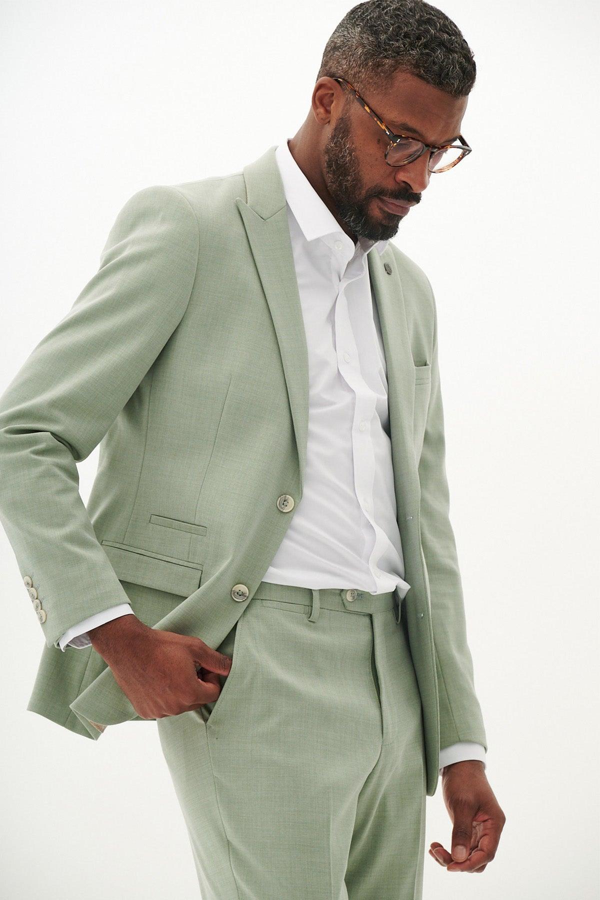 A man wearing glasses models the Marc Darcy FINLEY - Moss Green Two Piece Suit with a white shirt, looking down with one hand in his pocket against a plain white background.