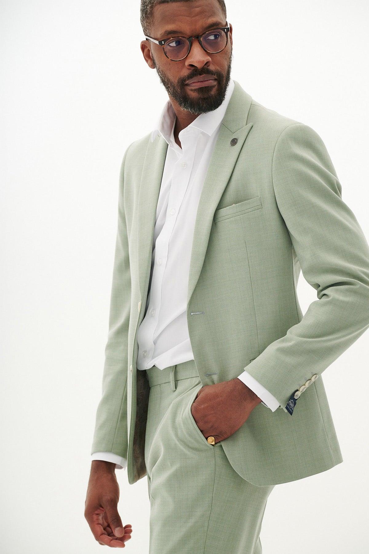 A man in the Marc Darcy FINLEY - Moss Green Two Piece Suit and white shirt, sporting glasses and a beard, poses with his hand in his pocket—an ideal choice for a stylish summer wedding look.