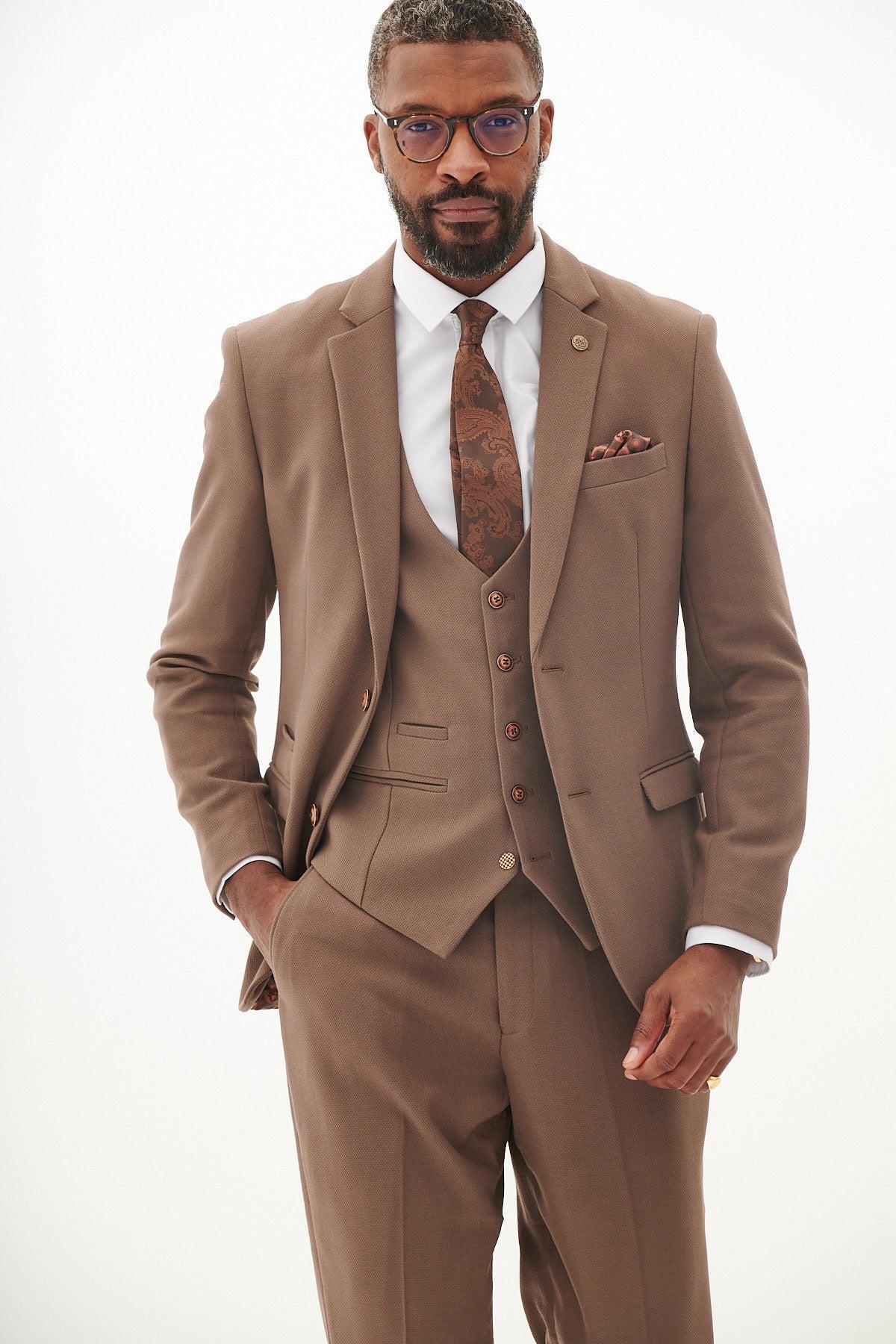 A man wears the marcdarcy HM5 - Tan Tailored Blazer with a matching vest, white shirt, patterned brown tie, and brown-framed glasses, standing with one hand in his pocket against a plain white background.