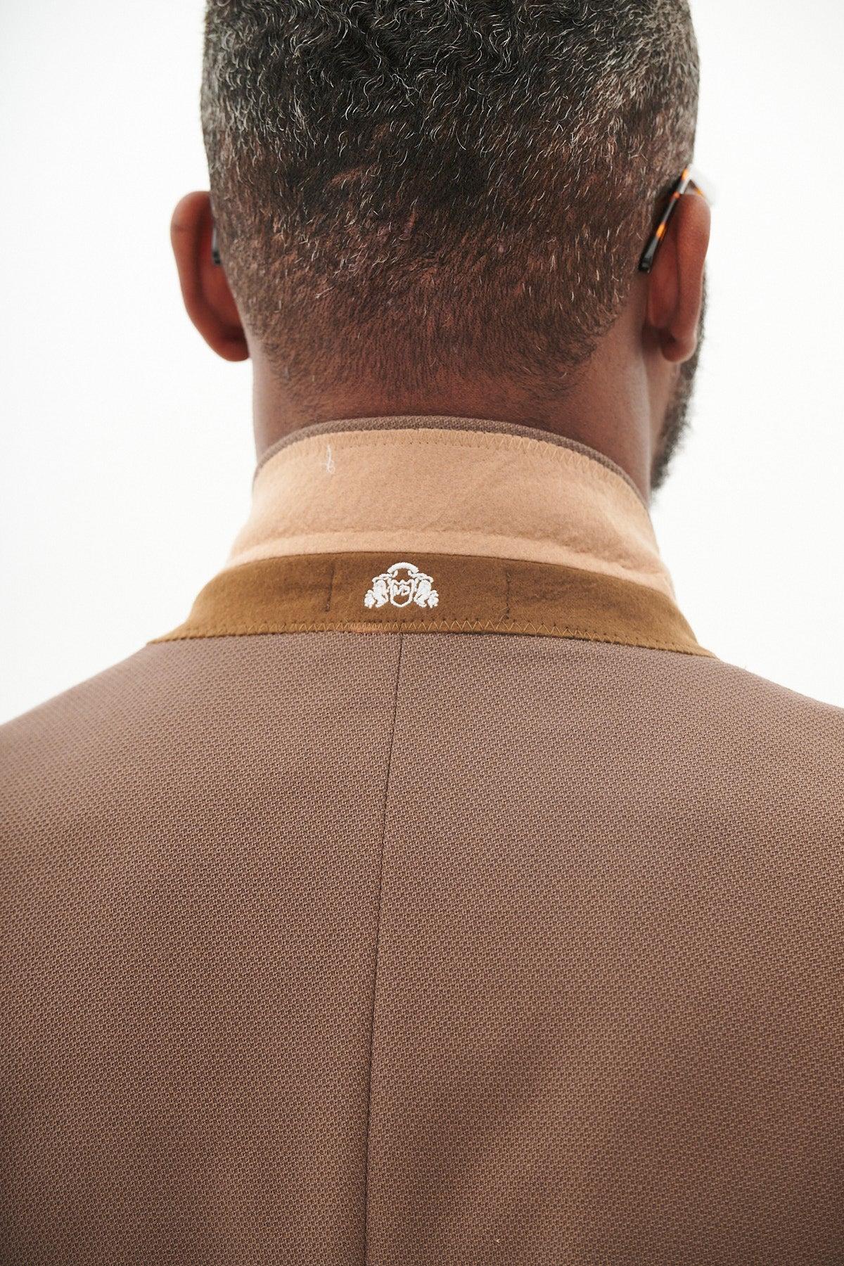 A close-up of a man’s back and neck wearing the Marc Darcy HM5 - Tan Blazer With HM5 Stone Waistcoat & Trousers, featuring a beige collar with a white emblem, short curly hair, and glasses visible on his ear.