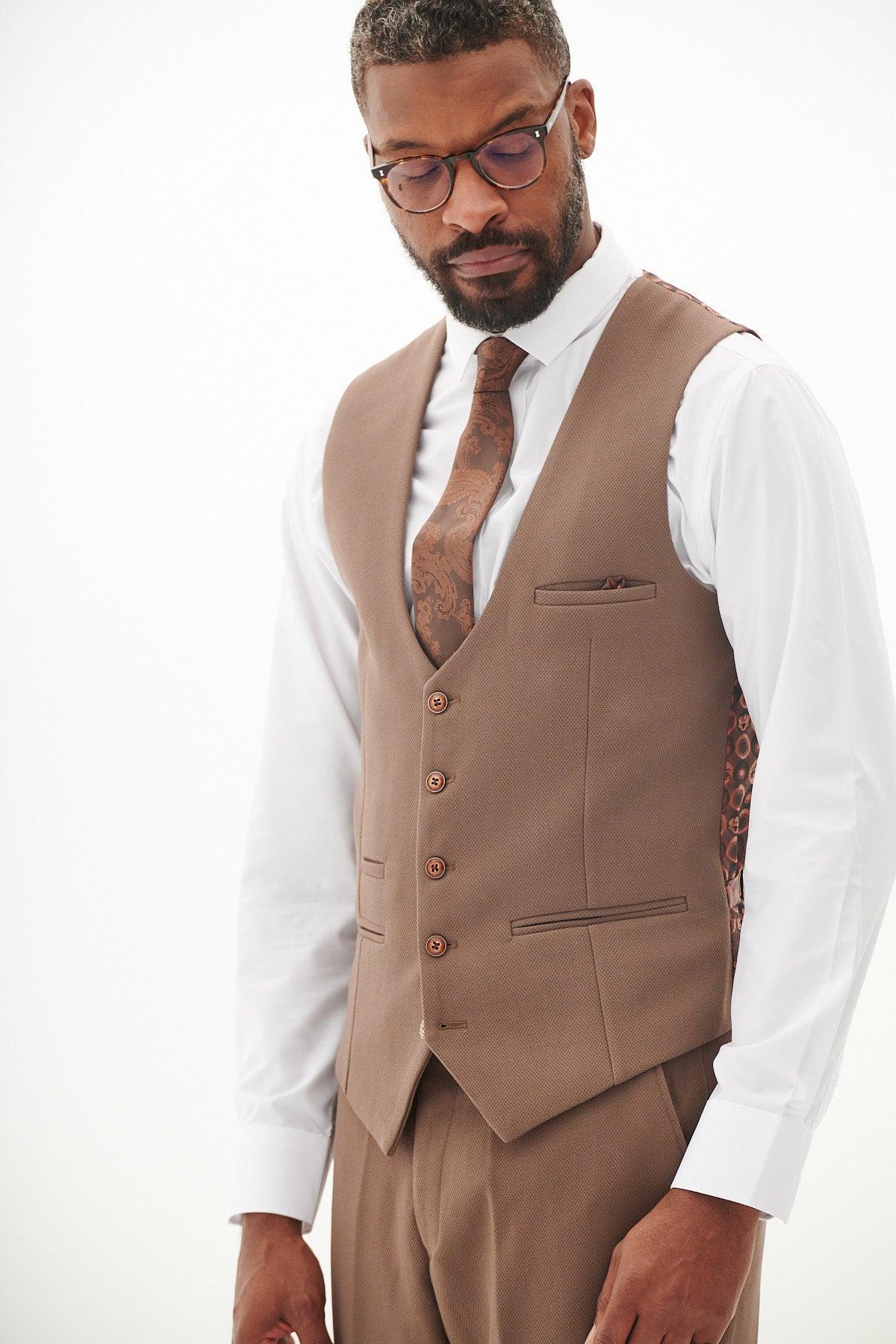 A Black man wears the marcdarcy HM5 Tan Single Breasted Waistcoat with a white shirt, brown paisley tie, and brown pants. His slim fit suit style is finished with glasses and a beard.