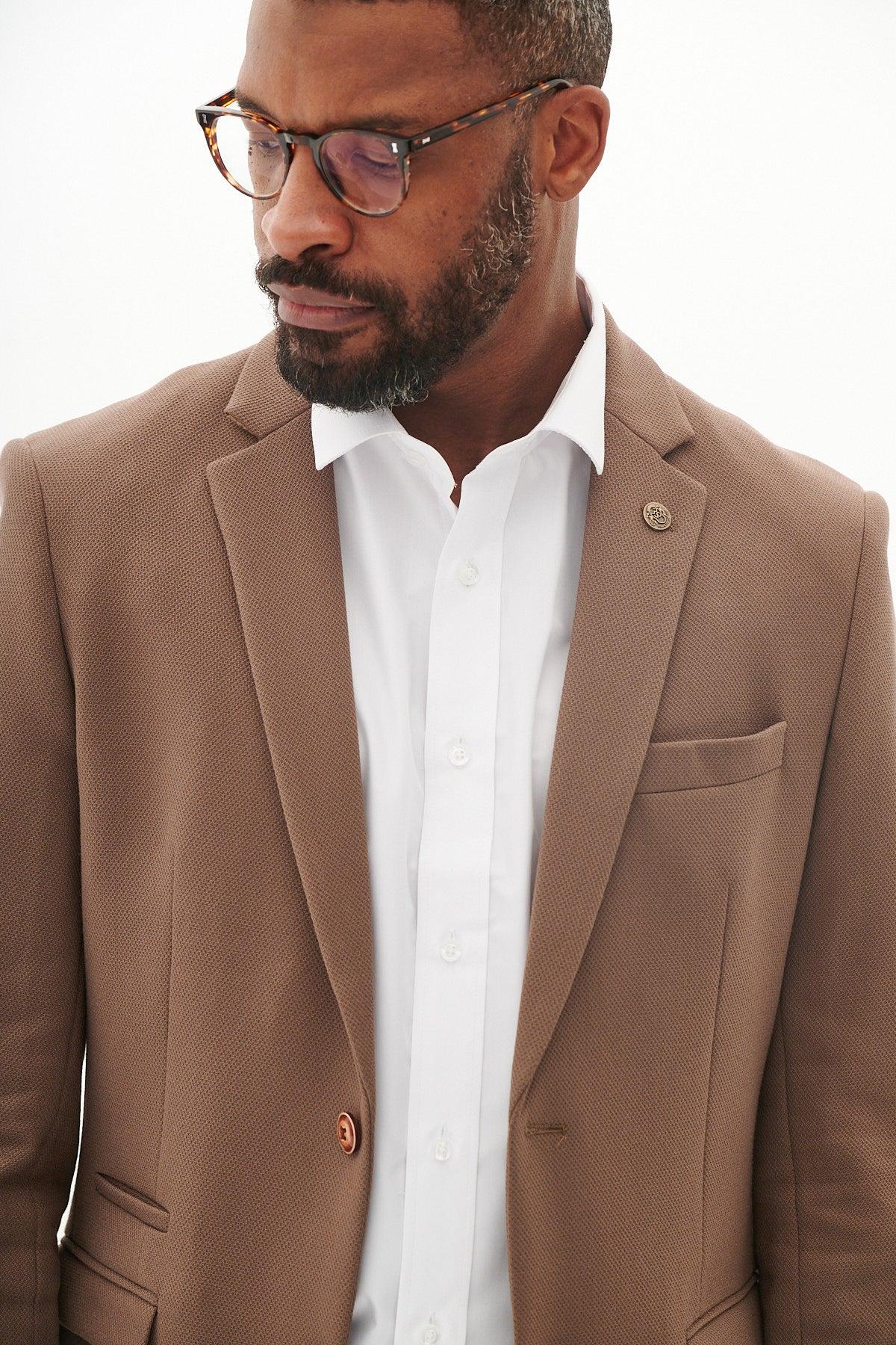 A man wears the Marc Darcy HM5 Tan Tailored Two Piece Suit with a white shirt and glasses, sporting a short beard and looking down against a plain light background.
