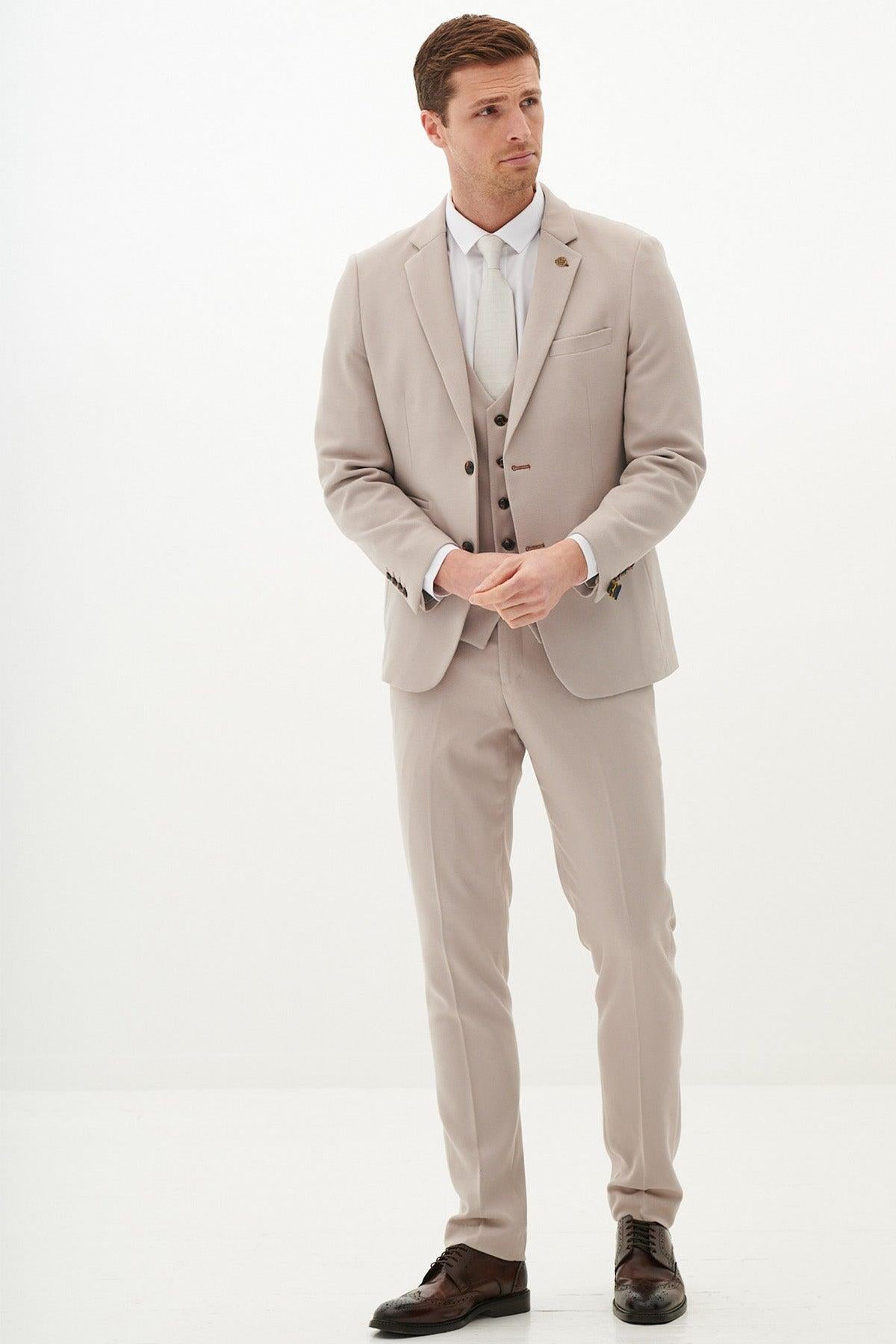 A man wearing the Marc Darcy Harry Maguire Wedding Suit—HM5 Stone Three Piece, with a white shirt and brown shoes—stands against a plain white background, looking slightly to the side with his hands clasped in front of him.