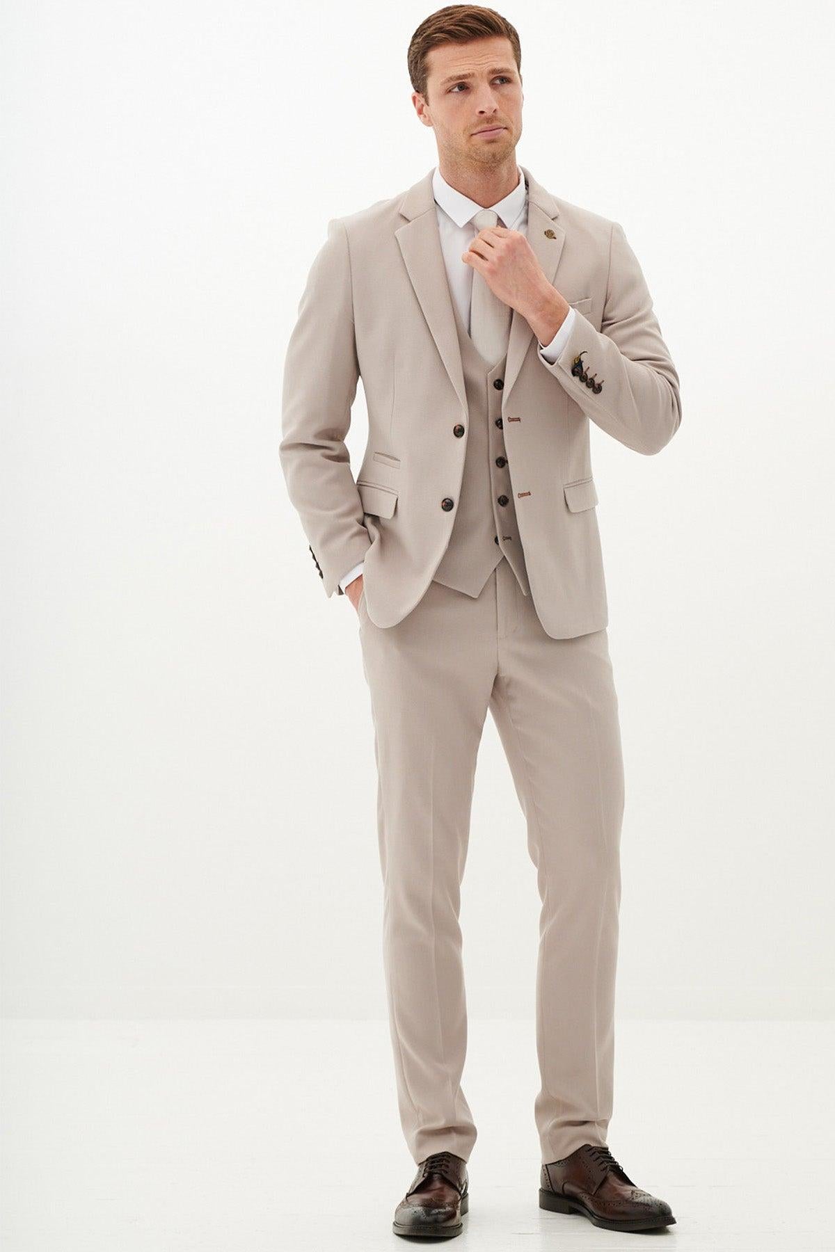 A man stands against a white background, wearing the Marc Darcy HM5 Stone Tailored Three Piece Suit with a white shirt and dark brown shoes. He adjusts his collar, channeling the refined style of a Harry Maguire wedding suit.