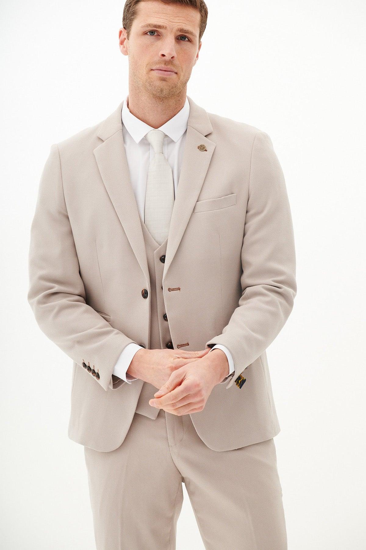A man wears the Marc Darcy HM5 Stone Tailored Three Piece Suit with a white shirt and matching tie, his hands clasped in front of him.