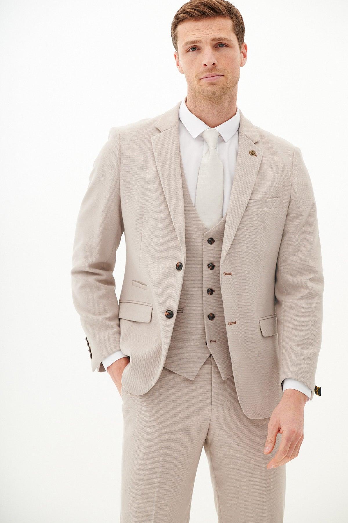 A man models the Marc Darcy Harry Maguire Wedding Suit HM5 Stone Three Piece, paired with a white shirt and tie, standing against a plain white background with one hand in his pocket and looking at the camera.