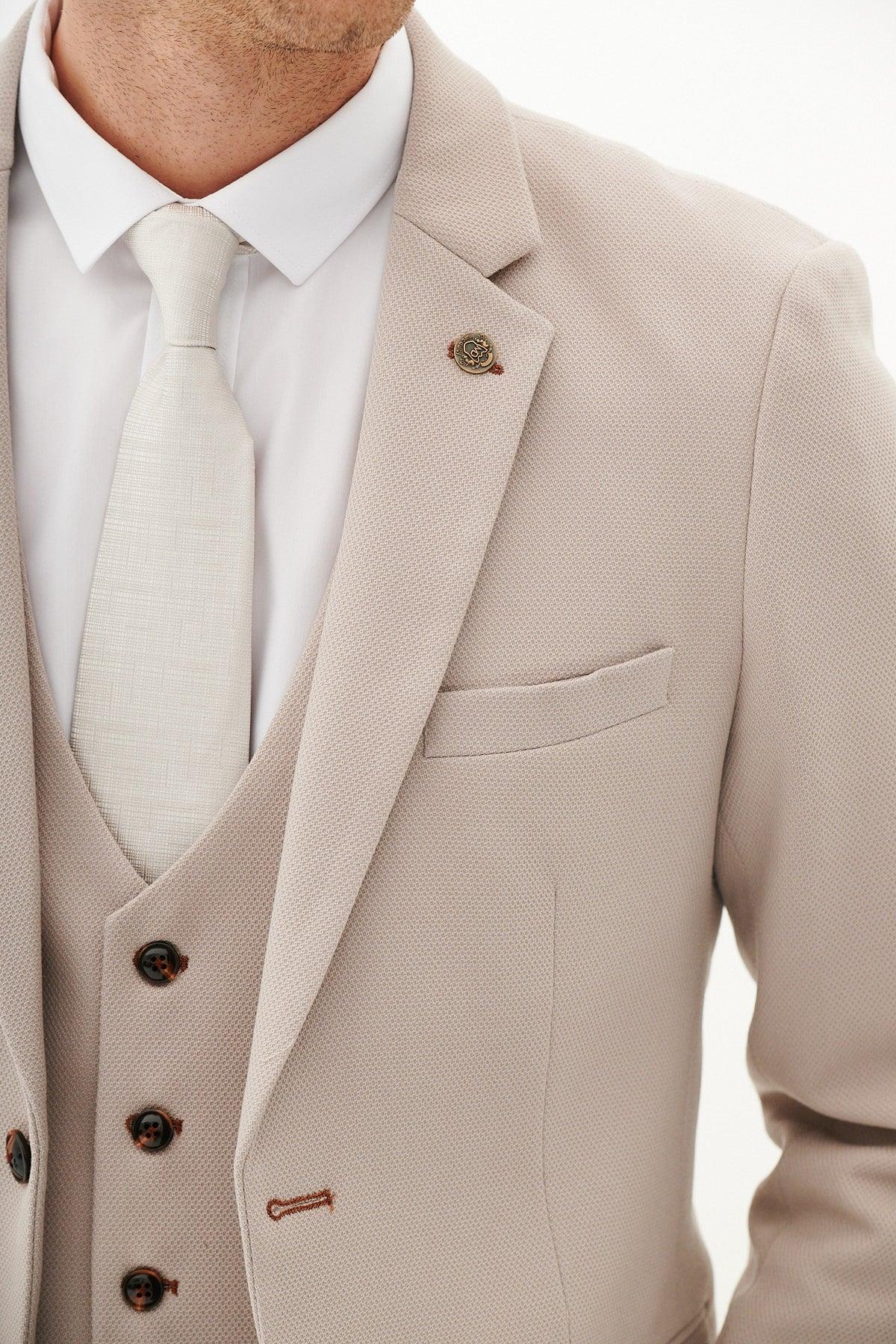 A person wears the marcdarcy HM5 - Stone Tailored Blazer, a slim-fit, light beige three-piece suit with brown buttons and a lapel pin, teamed with a white shirt and matching tie. Photo is cropped at the neck and midsection.