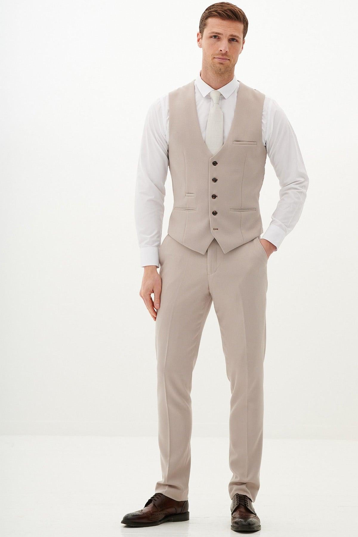 A man with short hair and a neutral expression wears the Marc Darcy HM5 - Stone Tailored Three Piece Suit with matching vest, white shirt, white tie, and dark brown shoes, standing against a plain white background.